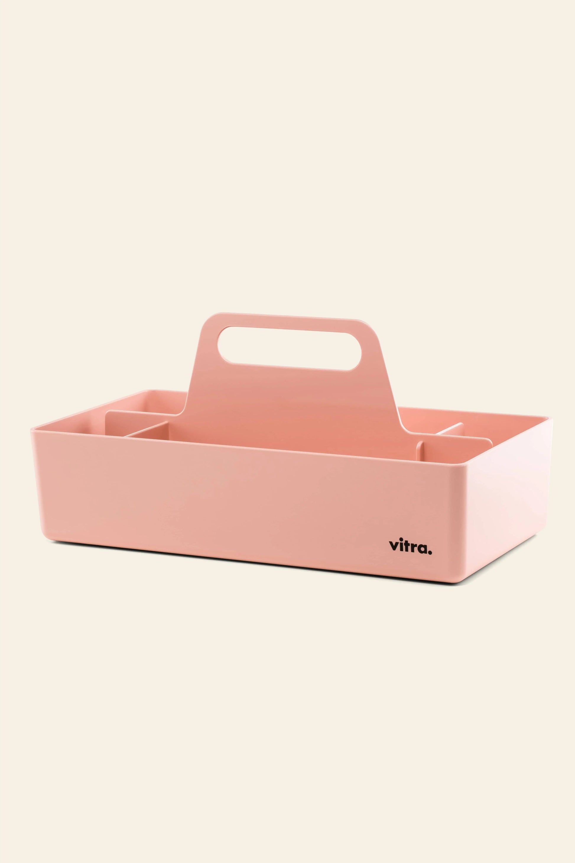 The Vitra Toolbox RE is a pink rectangular storage caddy made from recycled ABS plastic, with a central handle, multiple compartments, and the Vitra logo on the front corner. It’s shown against a light beige background.