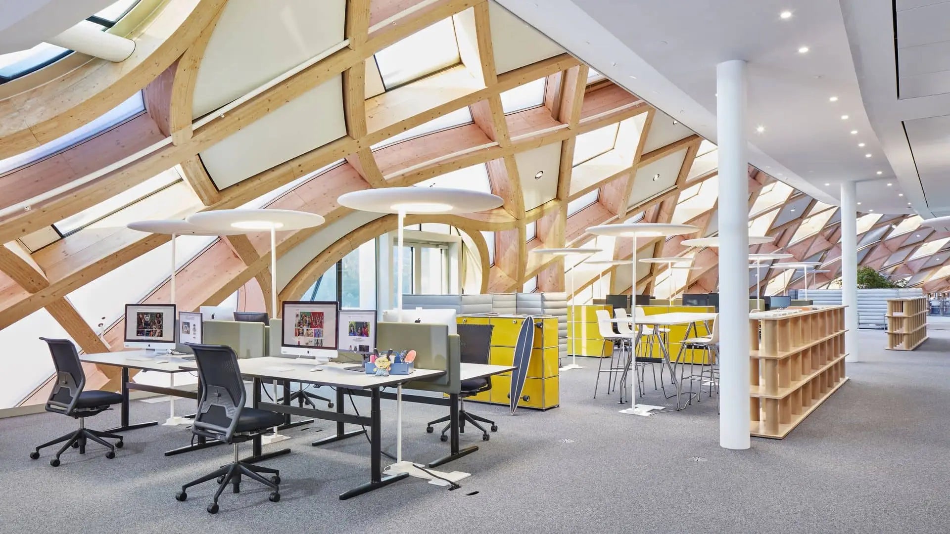 Modern office with curved wooden beams, large slanted windows, ergonomic chairs, yellow cabinets, high tables with stools, abundant natural light, and desks featuring Vitra’s Toolbox RE organizers made from recycled ABS plastic.