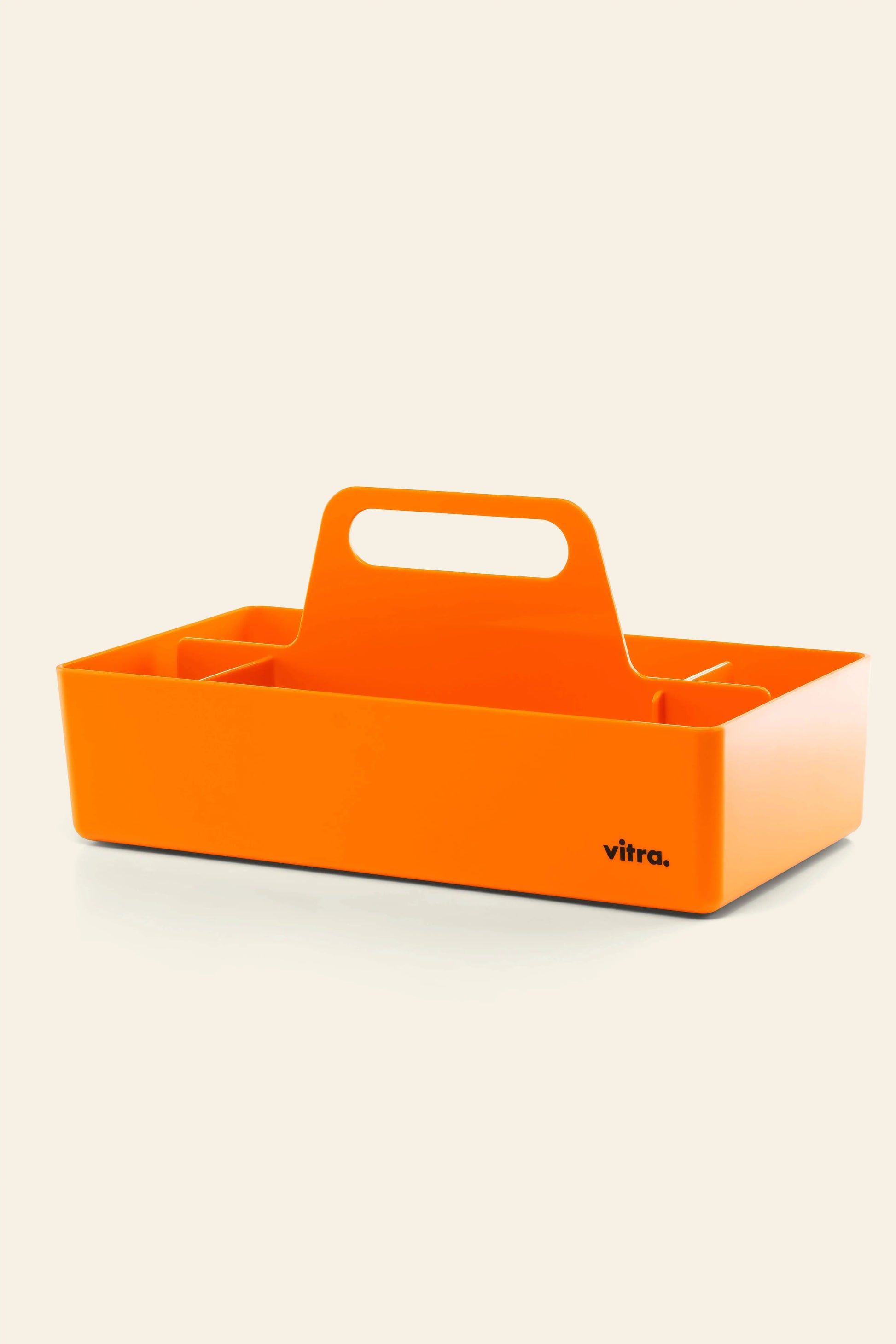 The Vitra Toolbox RE is a bright orange rectangular organizer made from recycled ABS plastic, featuring a central handle, multiple compartments, and “vitra.” branding on one side, shown against a plain light background.