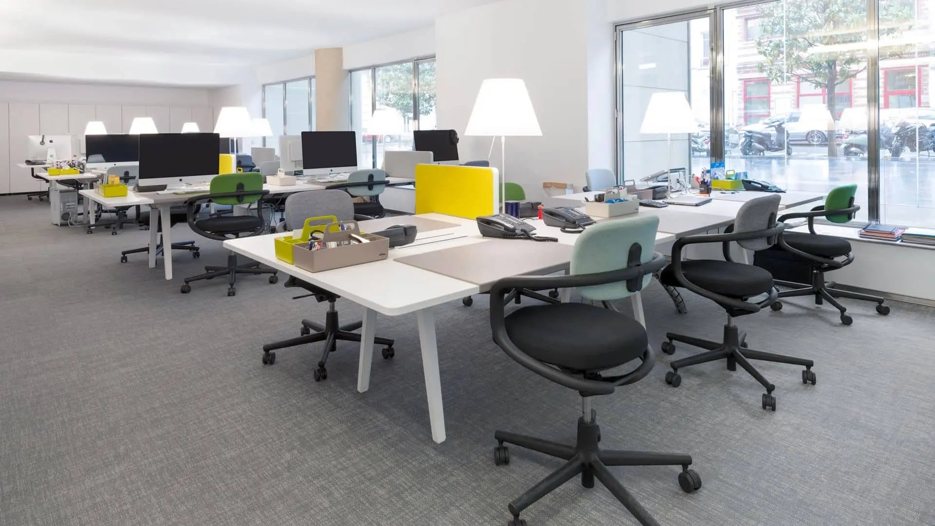 A modern open-plan office with empty desks, ergonomic chairs, Vitra computers, Vitra Toolbox RE organizers made from recycled ABS plastic, desk lamps, and large windows filling the clean, organized space with natural light.