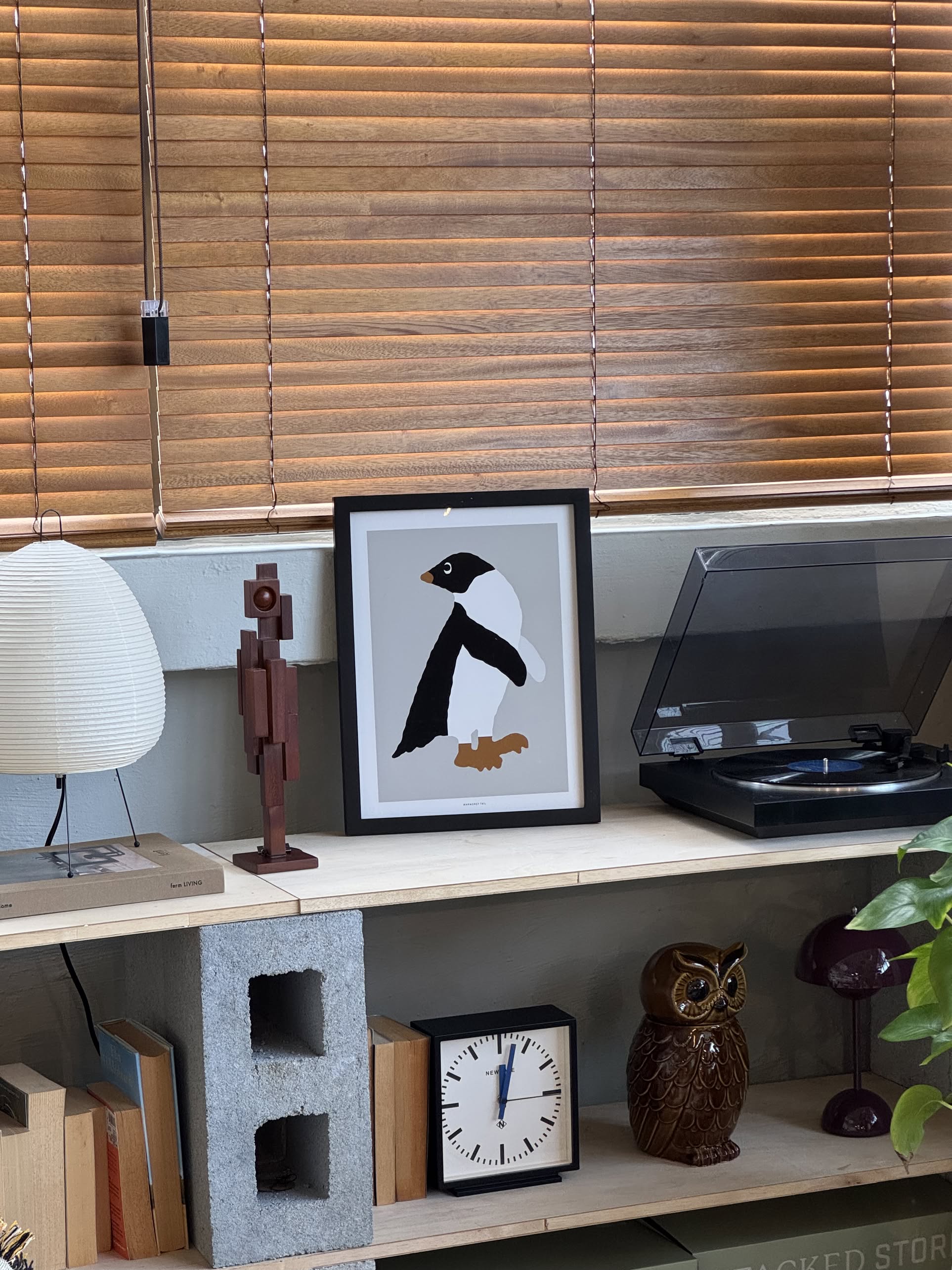 A shelf displays various items, including the WARMGREY TAIL Adelie Penguin Grey Poster (30x40) in a frame, a wooden owl figurine, a small analog clock, a record player, a lamp, a wooden figure, and a cinder block by the window.