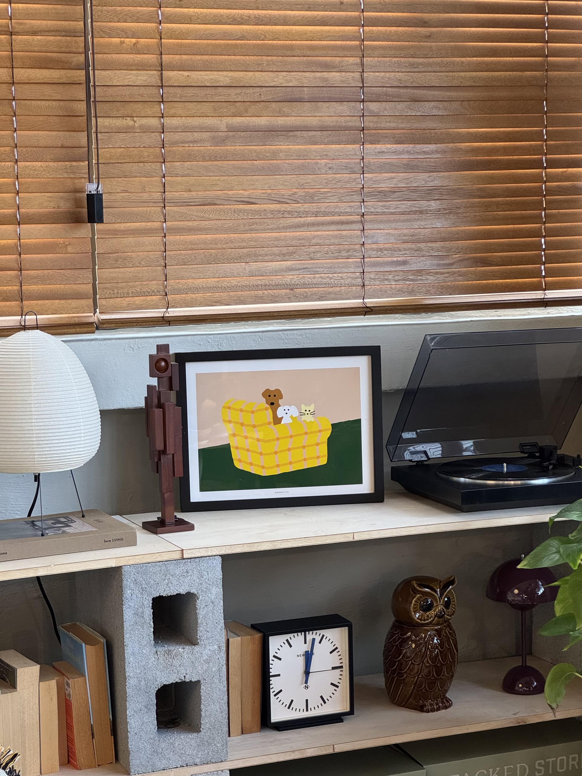 A shelf with the WARMGREY TAIL Armchair Dogs & Cat Poster (30x40), a wooden figure, lamp, vinyl record player, owl figurine, clock, and plant displayed underneath wooden blinds.