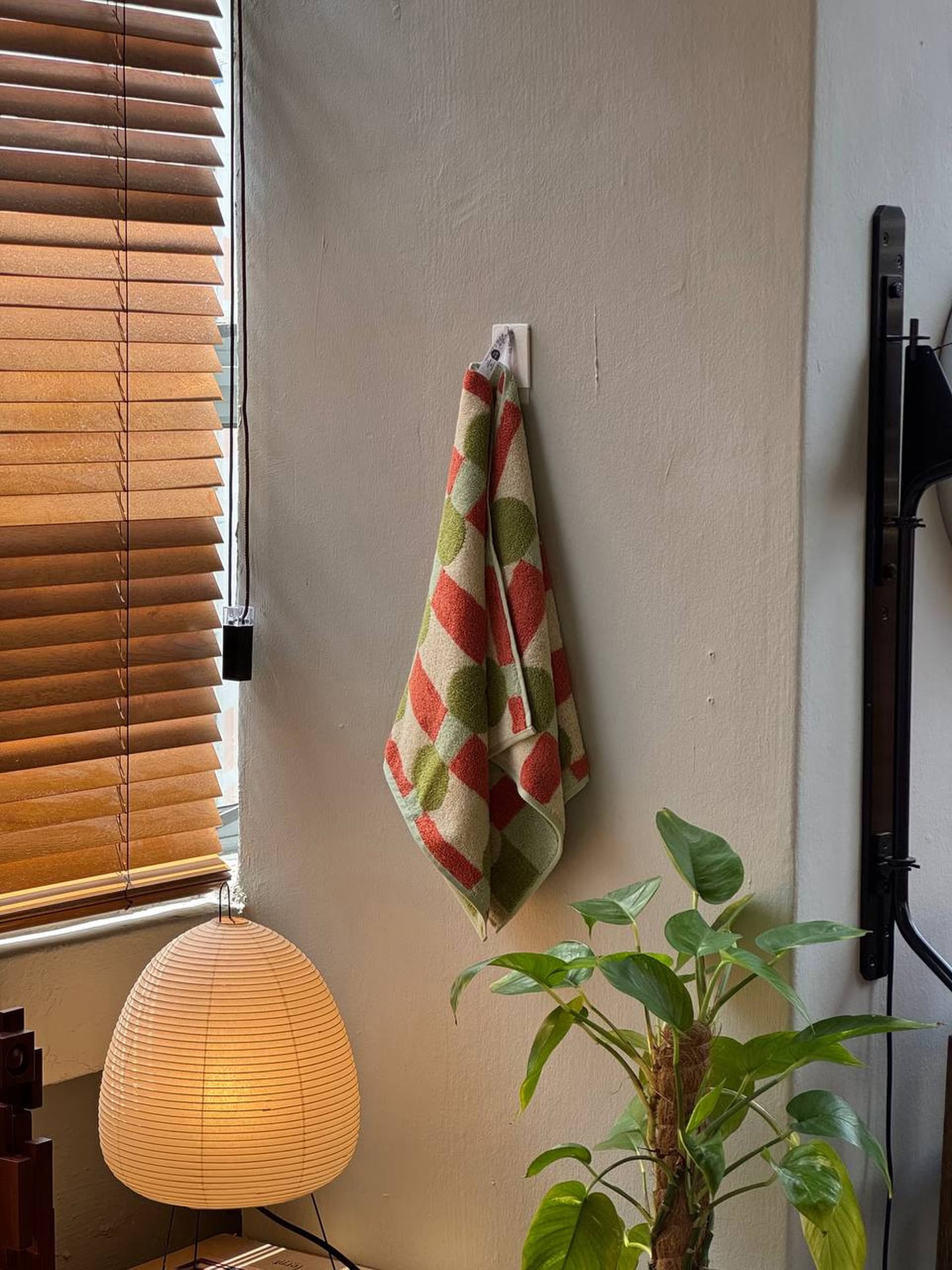 The WARMGREY TAIL Dot Check Face Towel by WARMGREY TAIL in green and orange hangs on a hook against a white wall near wooden blinds, a paper lamp, and a leafy plant as sunlight brightens the cozy, well-decorated space.