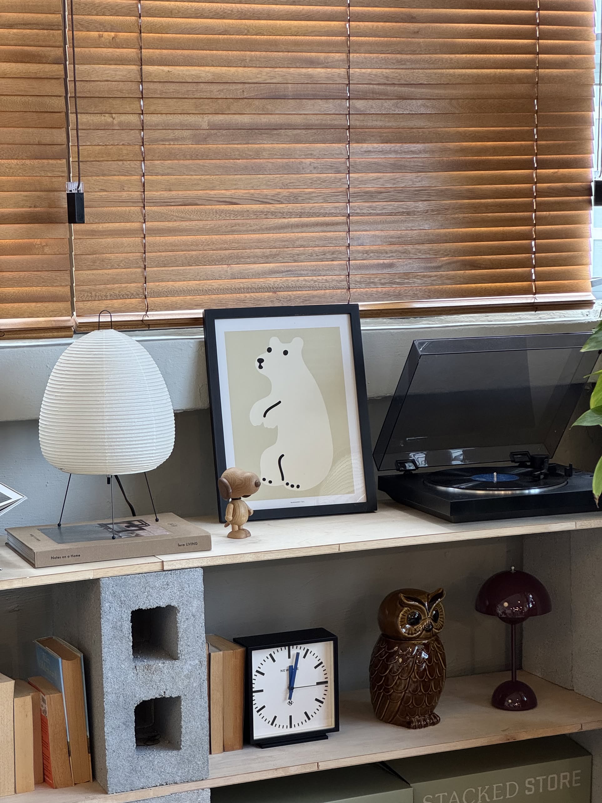 A bookshelf holds books, WARMGREY TAIL Huggy Bear Cream Poster (30x40), a teddy bear, an owl figurine, a clock, white and purple lamps. A record player sits on top, with wooden blinds covering the window behind.