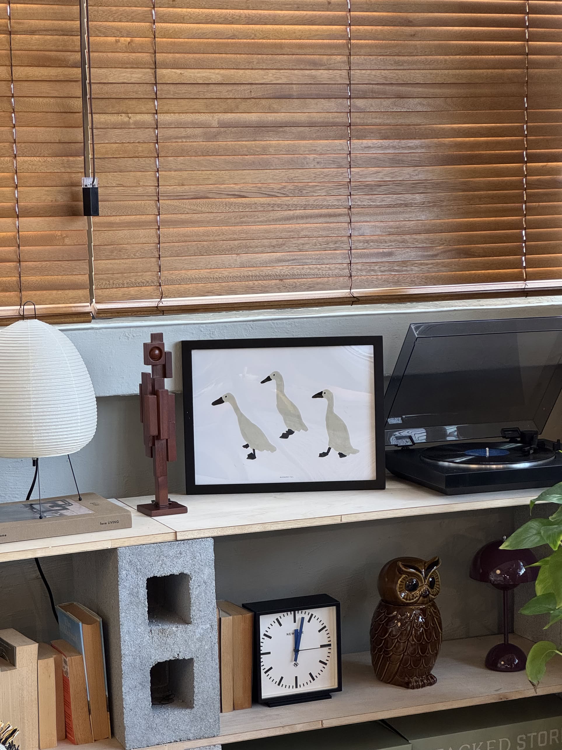 A shelf displays the WARMGREY TAIL Indian Runner Ducks Poster (30x40), a clock, an owl figurine, a wooden figure, a turntable, a lamp, and books. Minimal-style wooden blinds cover the window above.