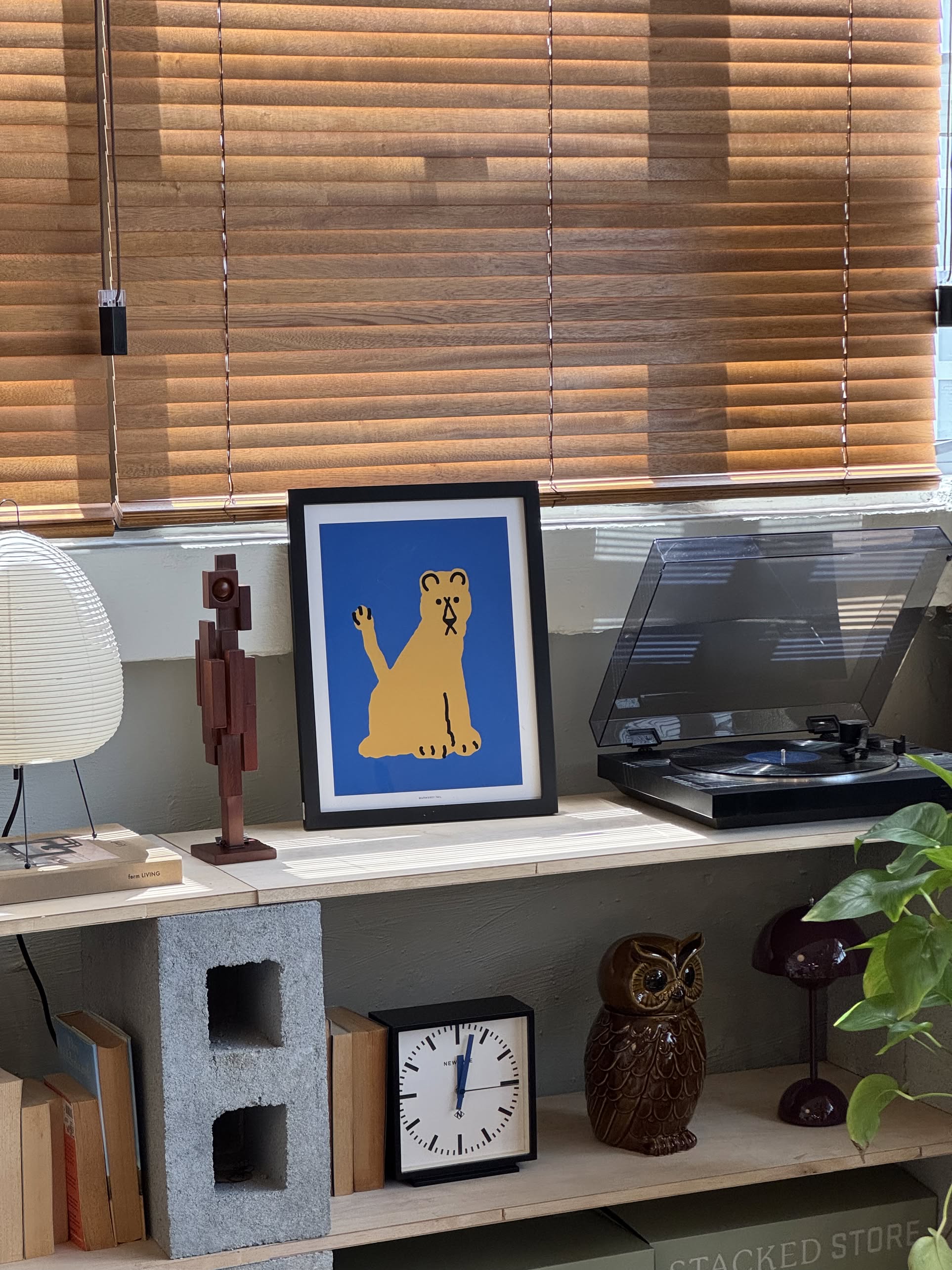 The WARMGREY TAIL Lioness Brown Poster (30x40), featuring minimalist lines in earthy tones, is framed and displayed on a wooden shelf beside a record player, owl figurine, table lamp, books, clock, and brown blinds.