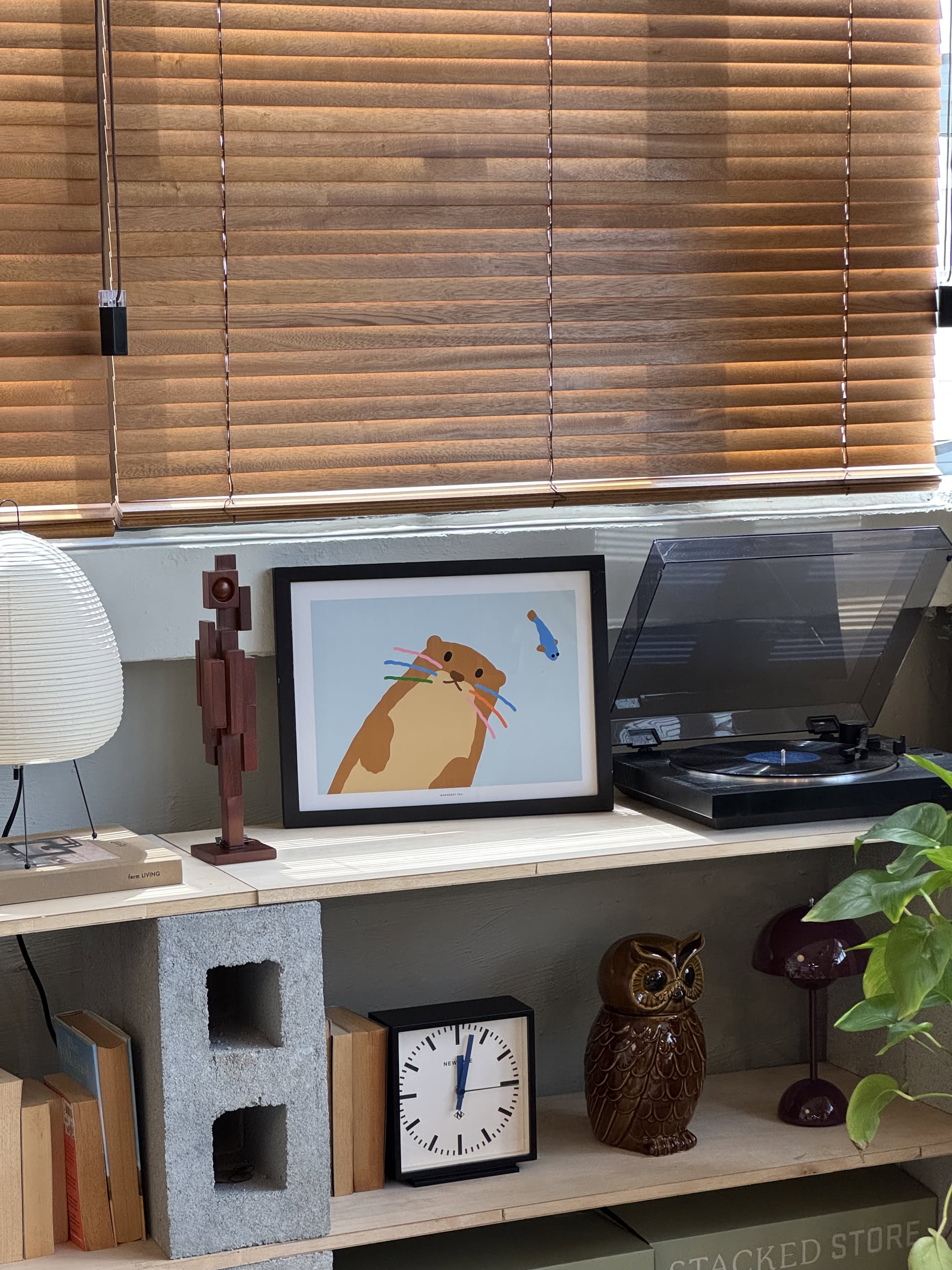A shelf with books, a wooden figurine, retro clock, ceramic owl, record player, green plant, and the WARMGREY TAIL Otter Light Blue Poster (30x40) by WARMGREY TAIL. Wooden blinds cover the window above a light blue wall.