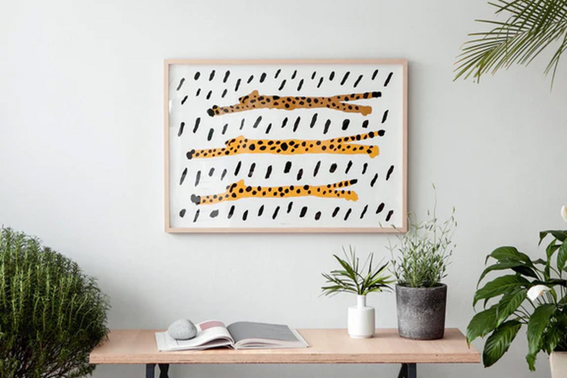 A wooden table with potted plants and an open book sits against a white wall, topped by the WARMGREY TAIL “Run Cheetah Run!” Poster (30x40), a minimalist artwork of three abstract cheetahs by WARMGREY TAIL.