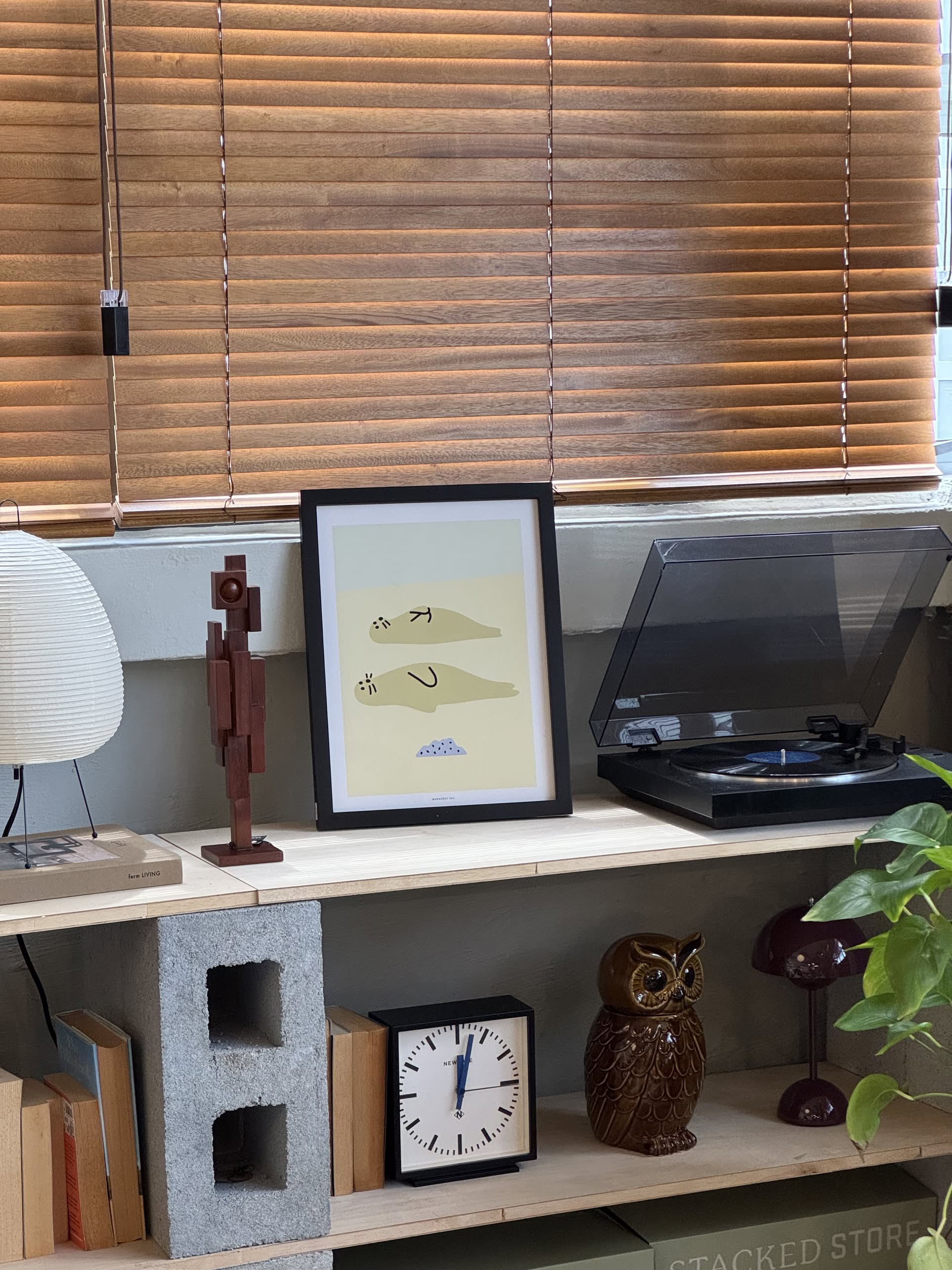 A shelf showcases the WARMGREY TAIL Seal Friends Poster (30x40), a sculpted figure, a record player, an owl figurine, a clock, plant, books, and decor blocks—all beneath wooden blinds that capture coastal charm.