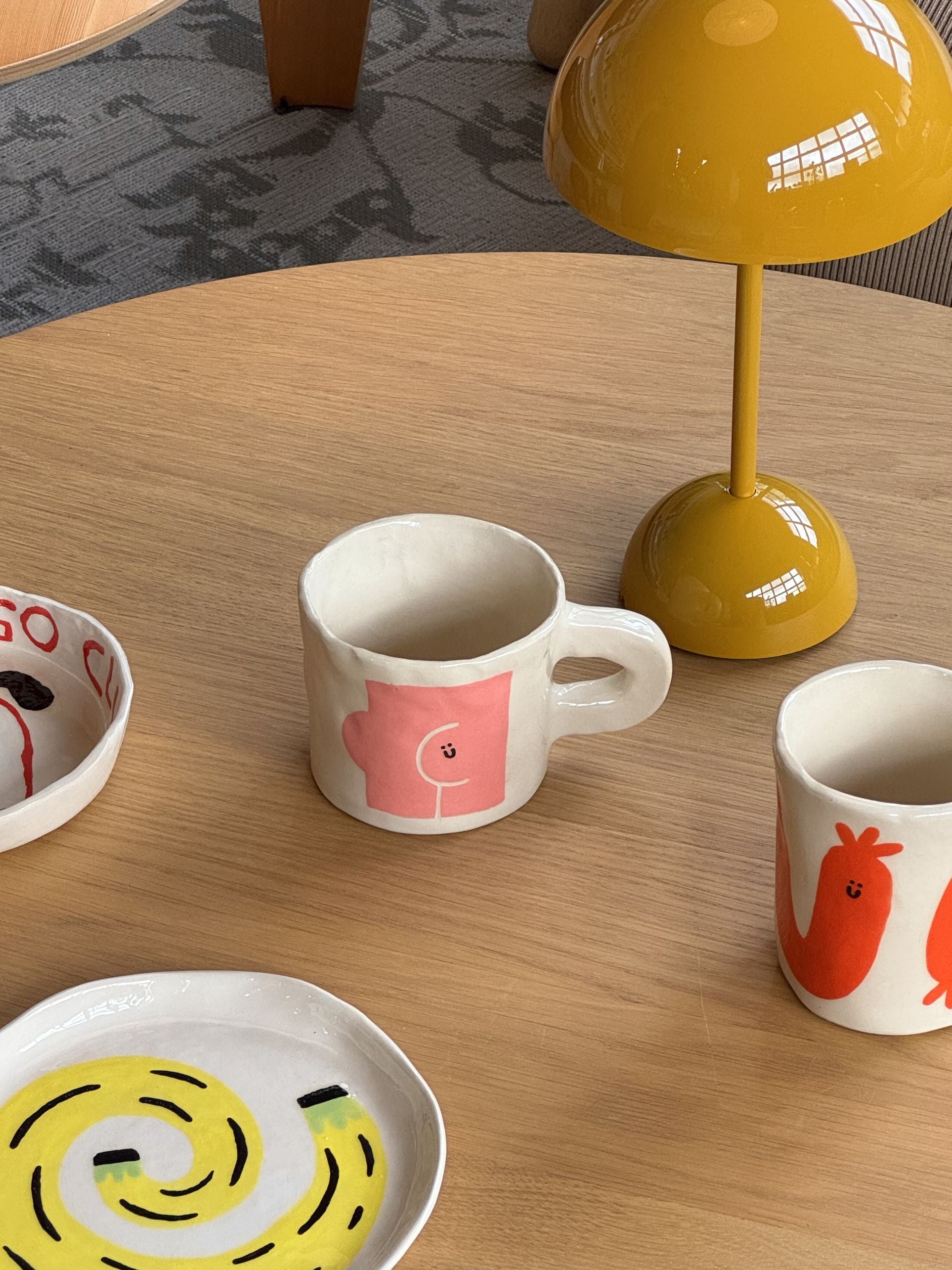 Wobbly Studios ceramic mugs and plates with playful, colorful designs—including the humorous Wobbly Studio Butts Mug—sit on a light wooden table. A round mustard yellow lamp brightens this cozy, conversation-starting setting.