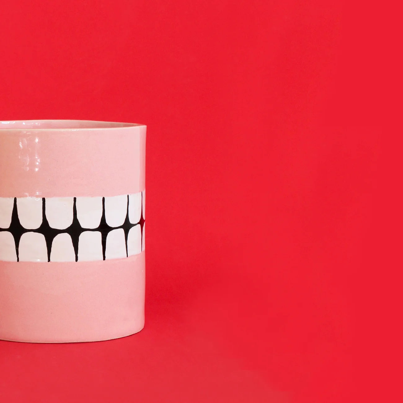 The Wobbly Studio Denture Pot by Wobbly Studio, a light pink ceramic cup with a black and white geometric design, makes a unique plant pot and is shown on the left against a bright red background.