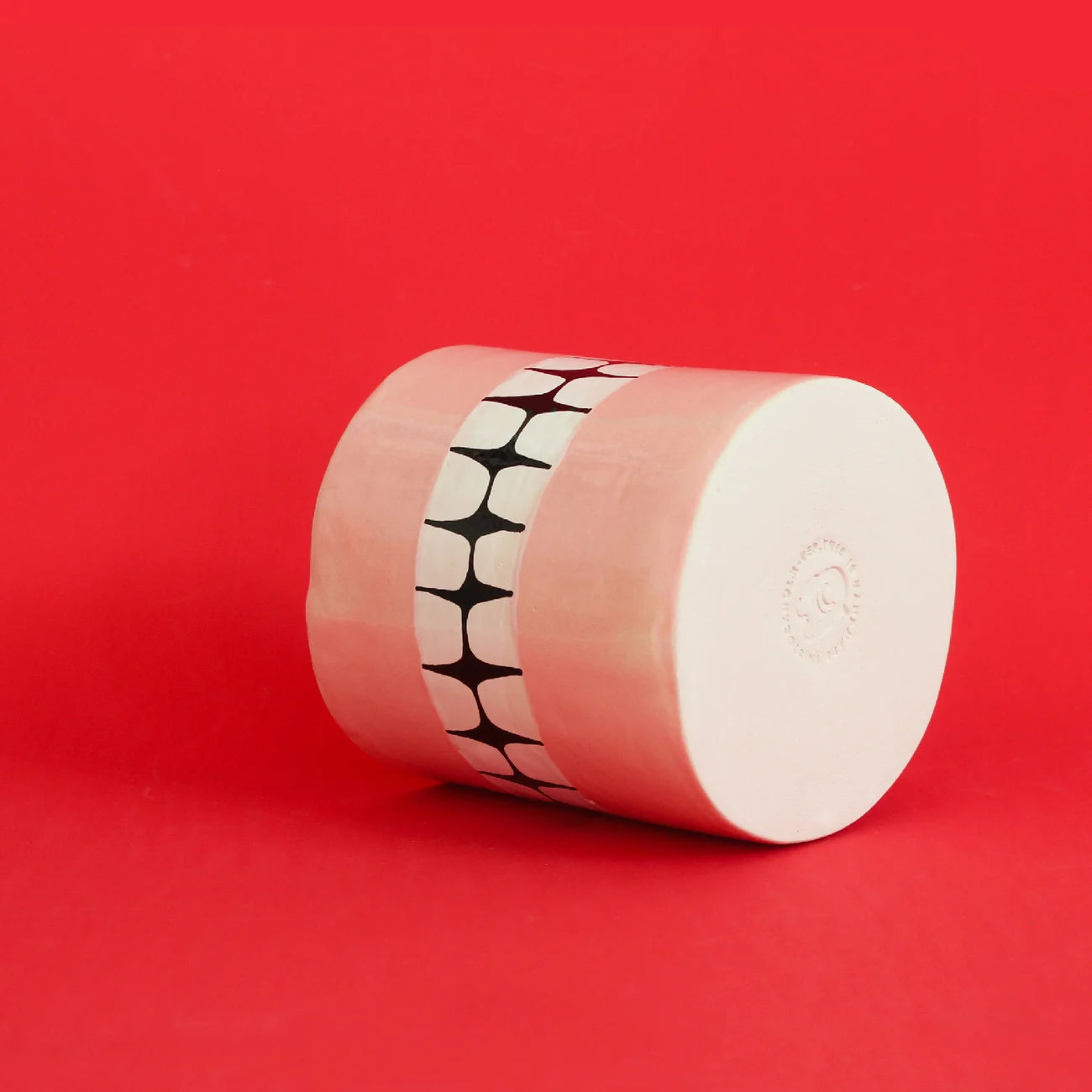The Wobbly Studio Denture Pot, a cylindrical ceramic piece with a pink and white finish and central black-and-white geometric band, rests on its side against a bright red background.