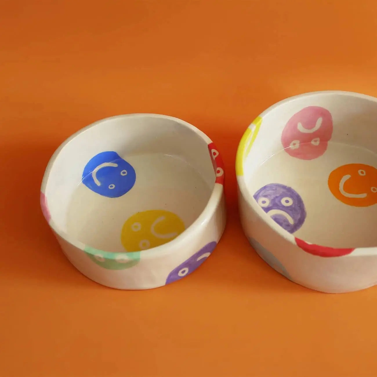 Two Wobbly Studio Doggo Bowl, Bigger by Wobbly Studio, each with multicolour faces and cheerful smiley designs inside and out, are displayed on an orange surface.
