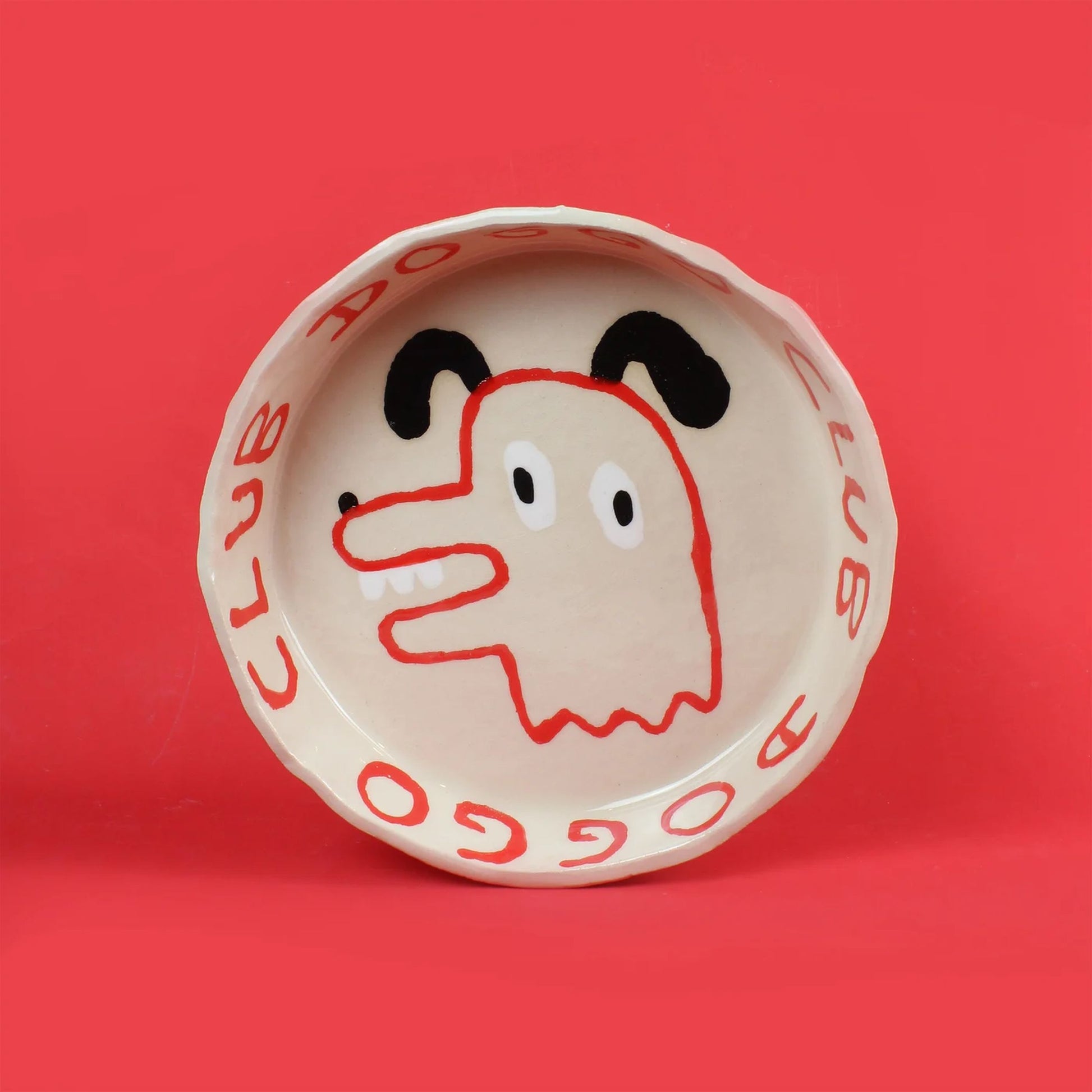 The Wobbly Studio Doggo Club Ti Chat Bowl is a ceramic plate featuring a cute dog face in red, black, and cream. “DOGGO CLUB” repeats on the rim in red over cream with a bold red background—perfect for your pet’s meals.