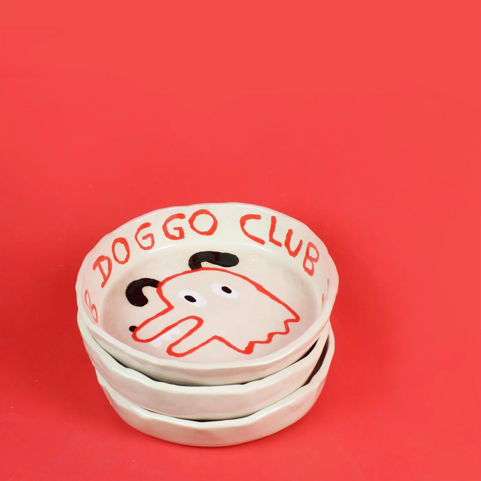 Three ceramic bowls are stacked on a red background. The top bowl is the Wobbly Studio Doggo Club Ti Chat Bowl by Wobbly Studio, featuring a cartoon dog face and DOGGO CLUB lettering—cute and functional for your pet’s meals.