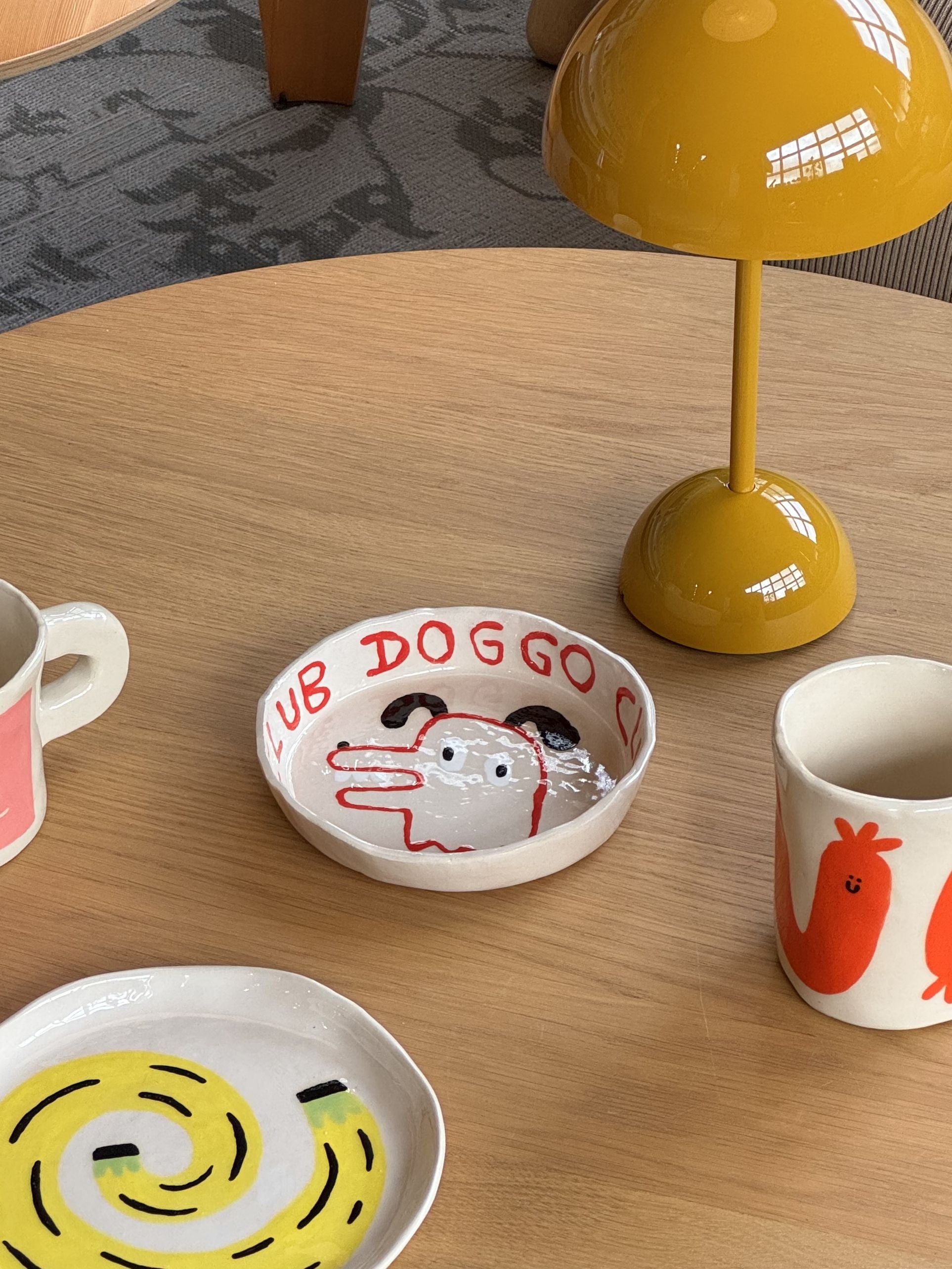 A round wooden table set with handmade ceramic dishes, including Wobbly Studios Doggo Club Ti Chat Bowl with a cute dog and LUB DOGGO text. Two mugs, a dish with a yellow swirl, and a small yellow lamp complete the cozy scene.