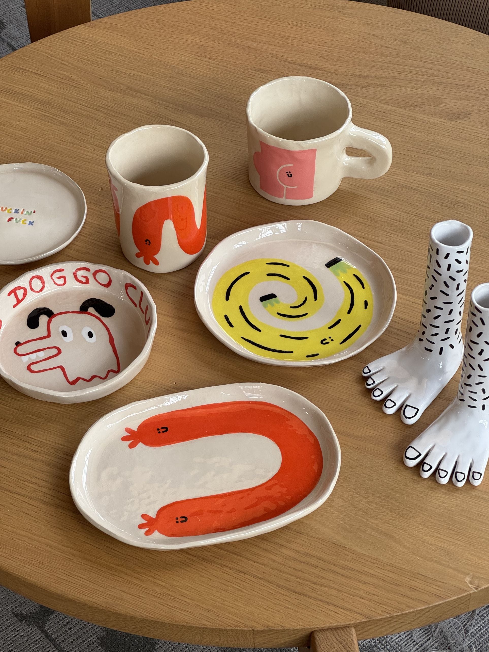 A selection of whimsical Wobbly Studio ceramic dishes, cups, and figurines—including the Doggo Club Ti Chat Bowl for pets—and playful designs like foot-shaped pieces are displayed on a round wooden table.