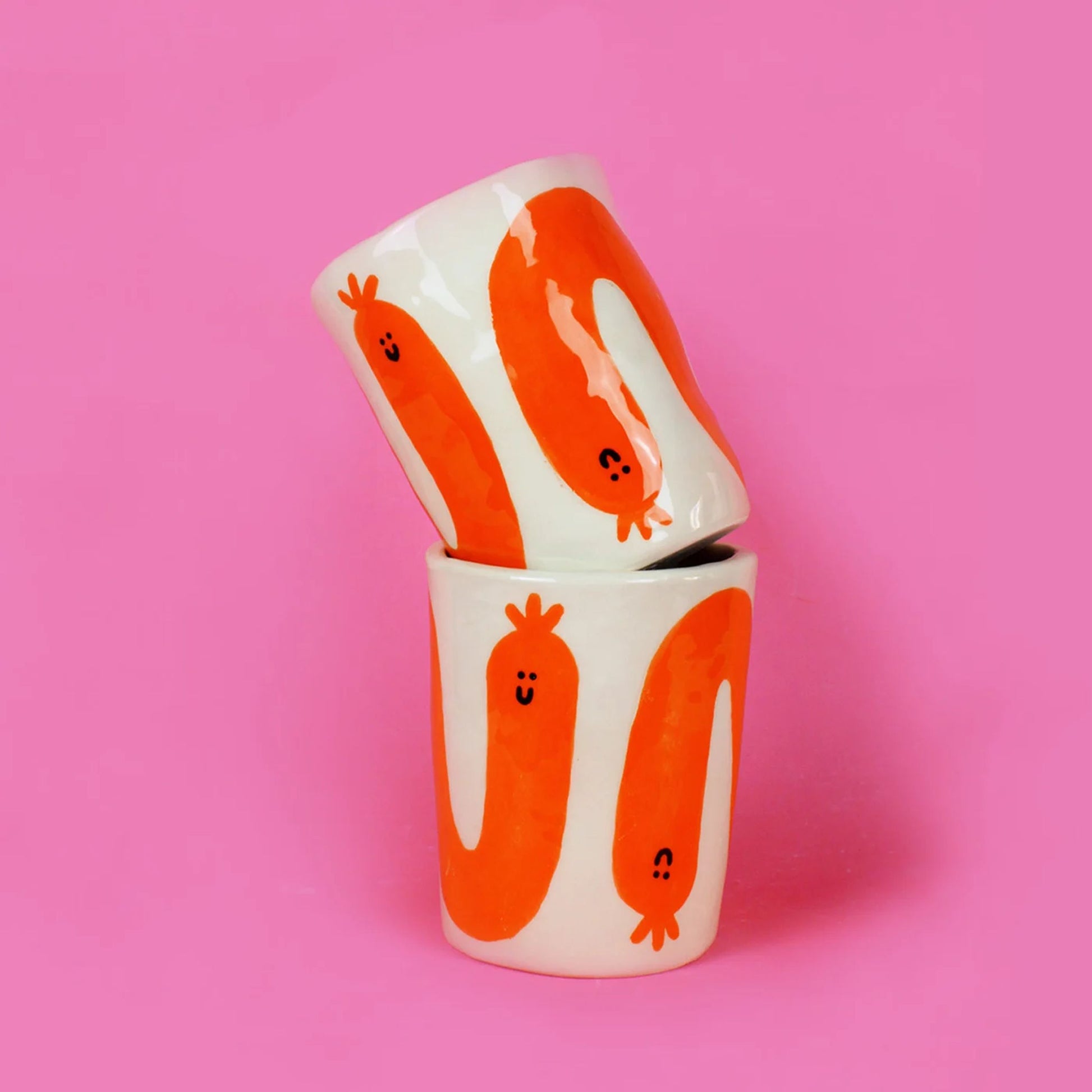 Two Wobbly Studio Happy Saucisse Tumblers with playful smiling worm designs are stacked against a solid pink background—a charming handmade earthenware addition from Wobbly Studio.