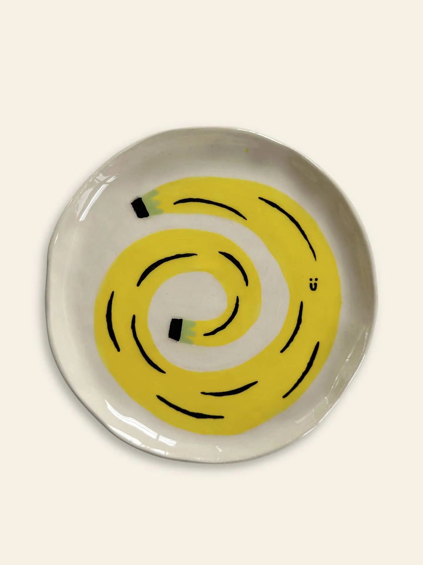 The Wobbly Studio Happy Things Plate - Banane is a ceramic plate with a cheerful, spiraled yellow pencil design, playful black lines, green-tipped ends, and a small black smiley face on an off-white background.