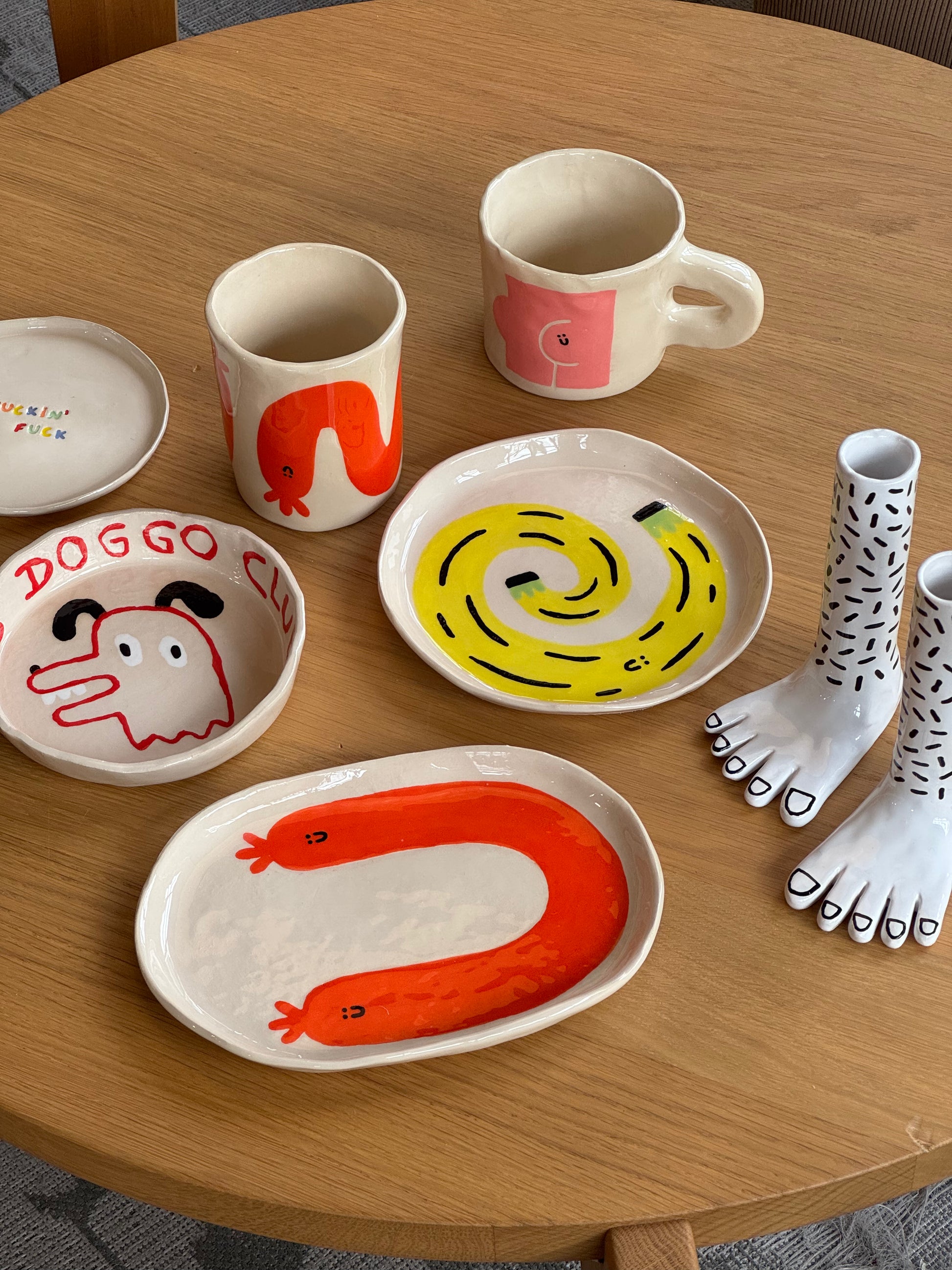 A set of whimsical ceramics on a wooden table, including mugs, bowls, and Wobbly Studio Happy Things Plate - Banane by Wobbly Studio—plates shaped like cheerful feet with playful patterns such as bananas and black dots.