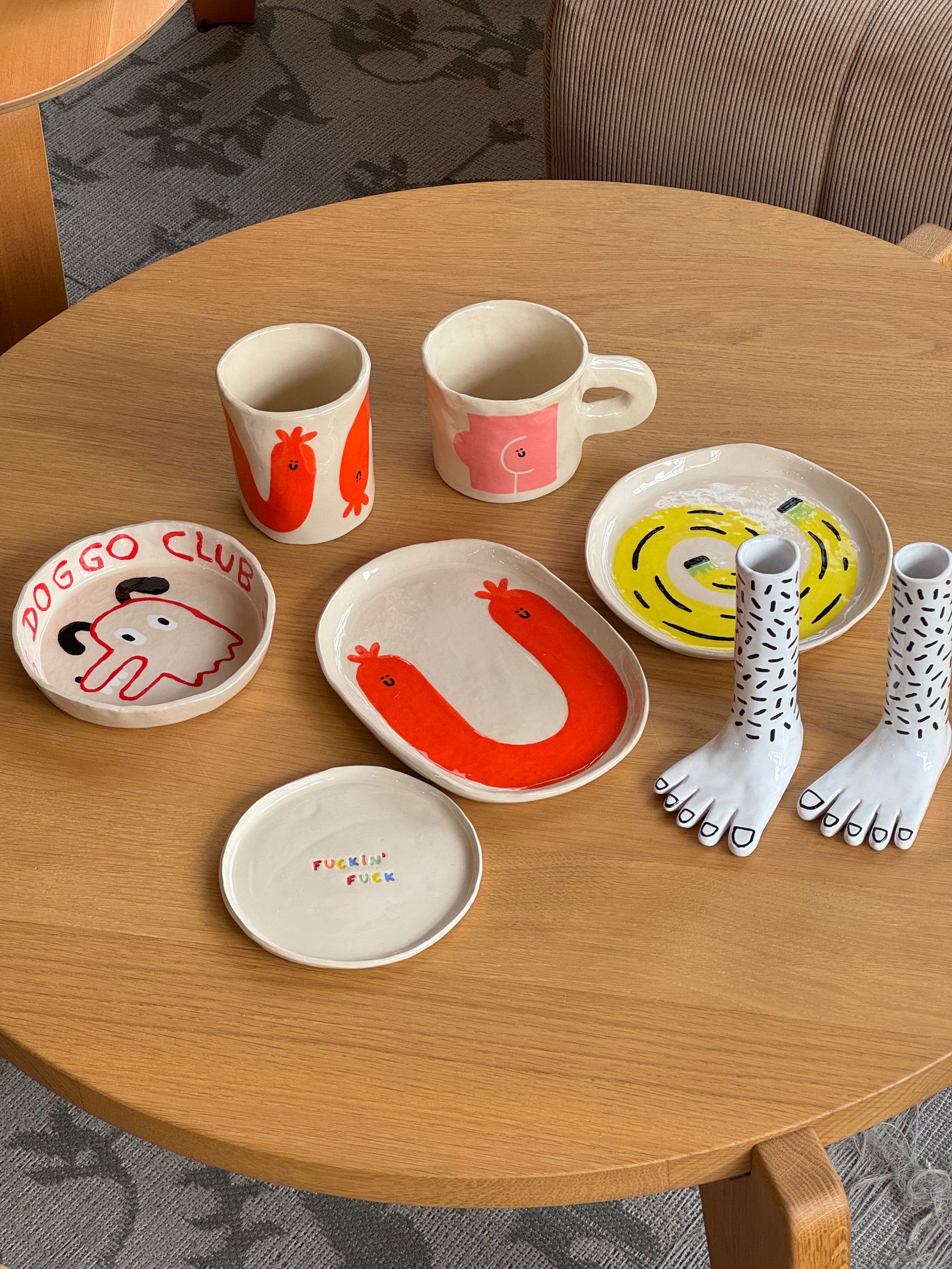 A round wooden table displays whimsical ceramic dishes, including the Wobbly Studio Happy Things Plate - Banane, featuring cheerful illustrations alongside cups, other plates, and foot-shaped sculptures with painted black nails.