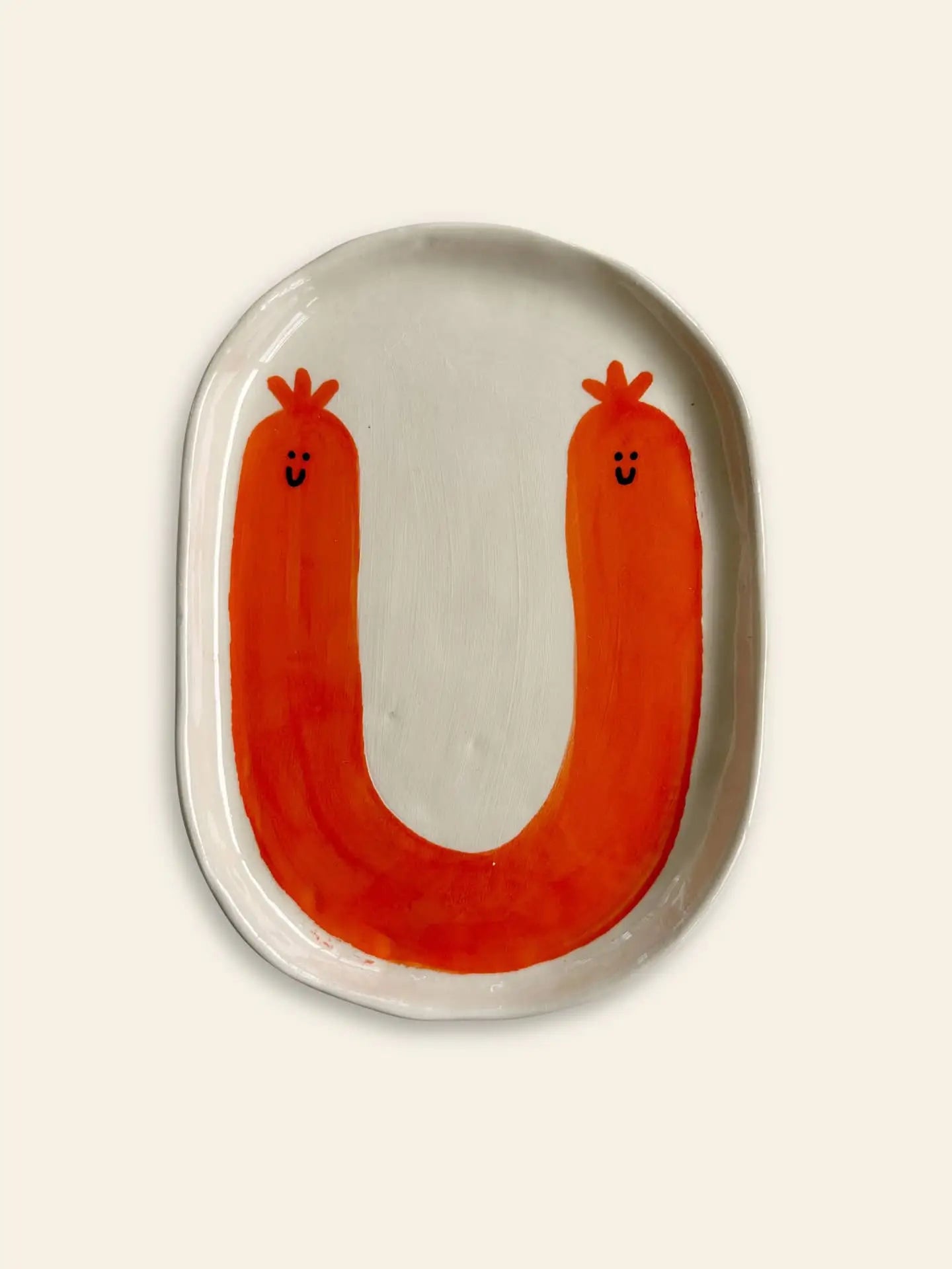 The Wobbly Studio Happy Things Plate - Saucisse is a white ceramic plate featuring a large orange U-shape with smiling faces and crowns, resembling playful sausages in vibrant colors.