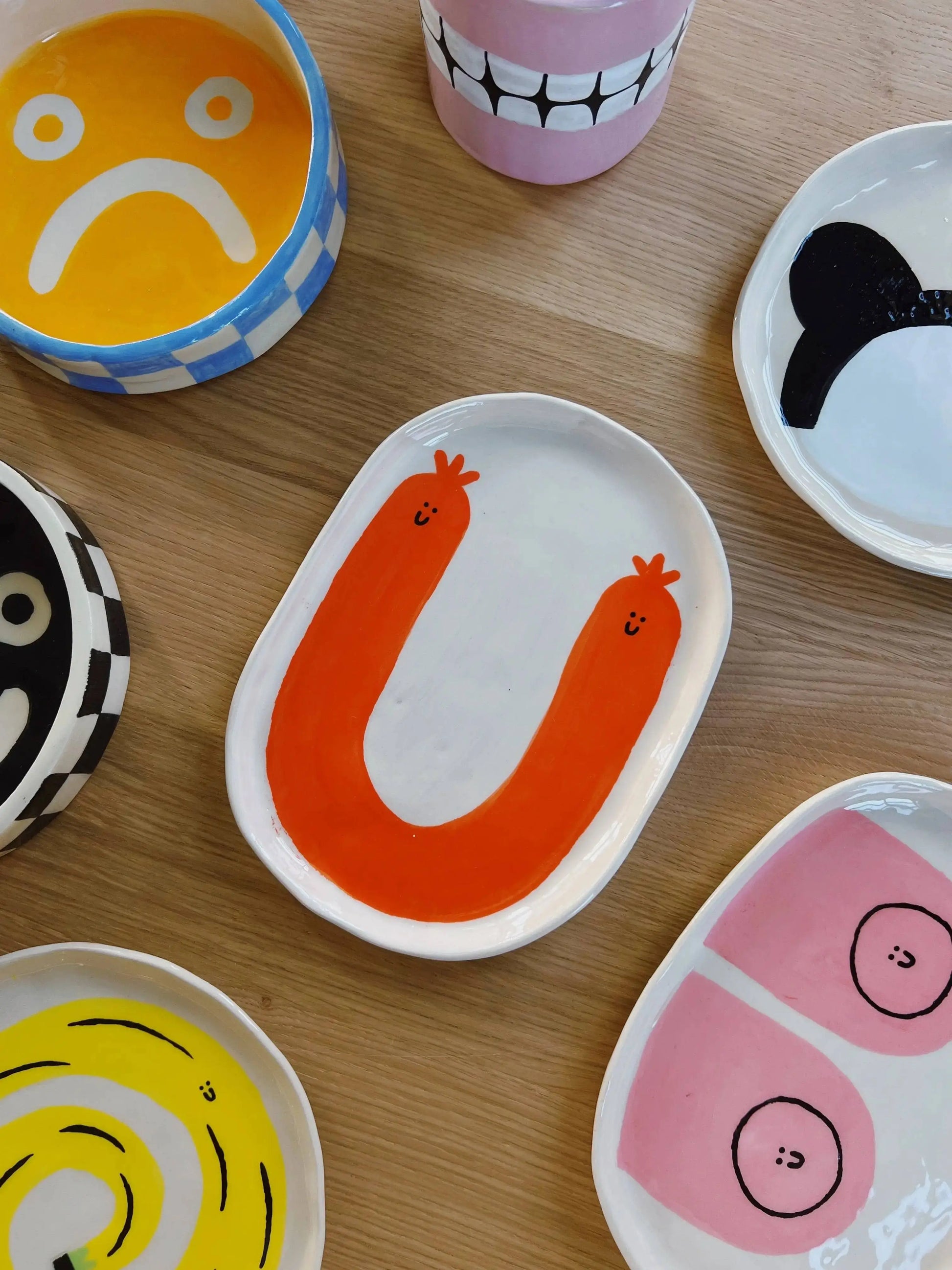 The Wobbly Studio Happy Things Plate - Saucisse by Wobbly Studio features colorful, hand-painted illustrations like a red U-shape with faces, a yellow sad face, black and white patterns, and a vibrant pink plate with two circles.