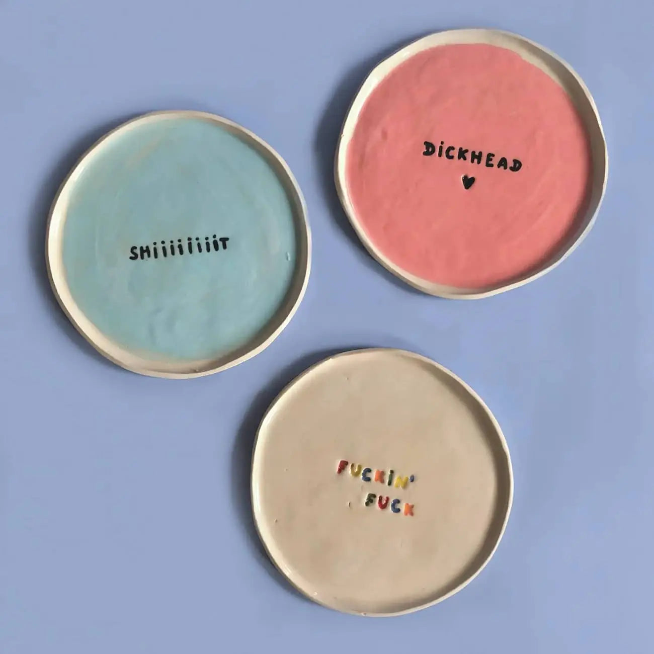 Three handcrafted Wobbly Studio mini plates—blue with shiiiiiit, pink with dickhead and a heart, and beige with colorful Fuckin Fuck lettering—add playful charm to any space. Features the Wobbly Studio Mini Plate - Fuckin Fuck.