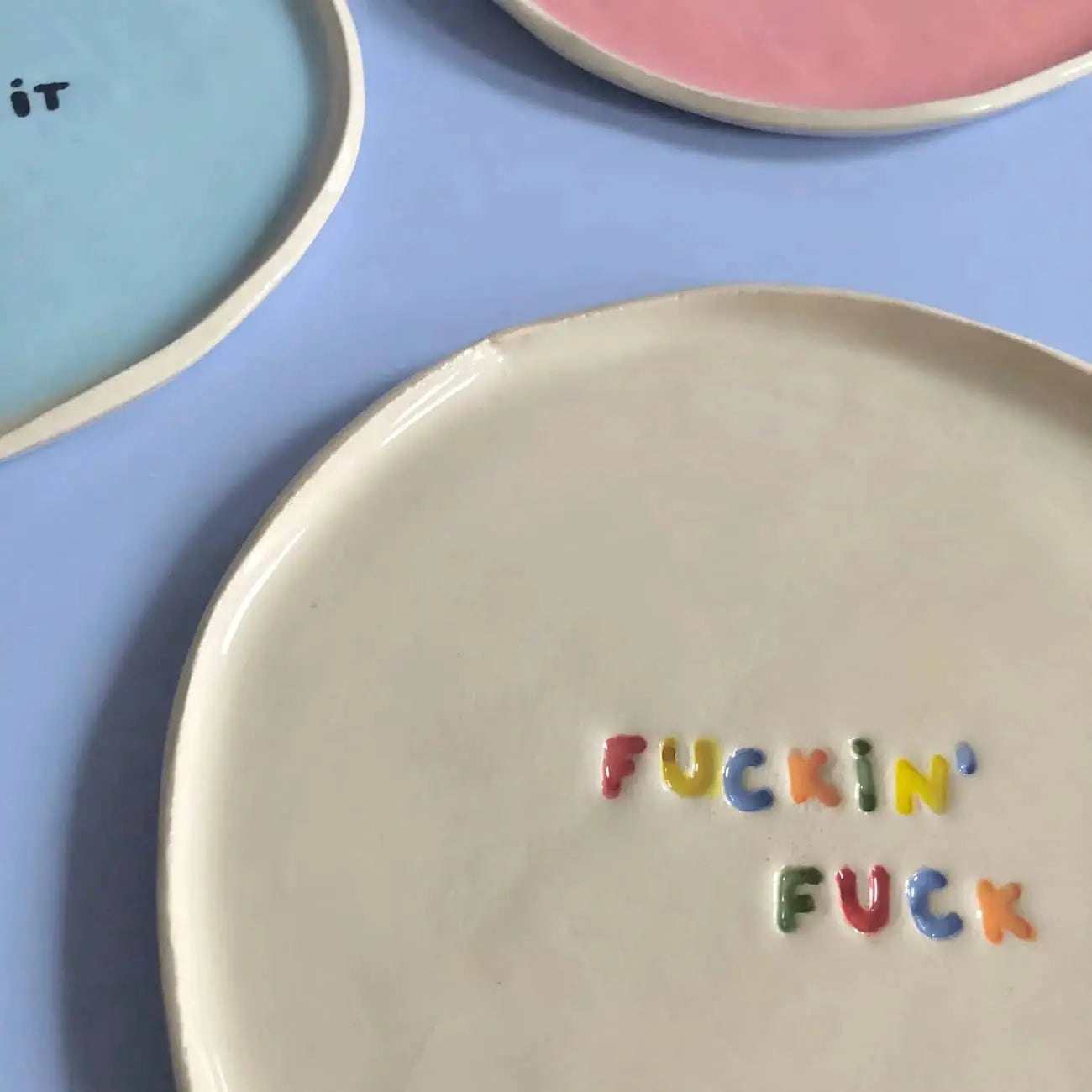 Three handcrafted Wobbly Studio mini plates rest on a pastel blue surface: one features colorful Fuckin Fuck text, another shows pastel letters, and the third is plain pink.
