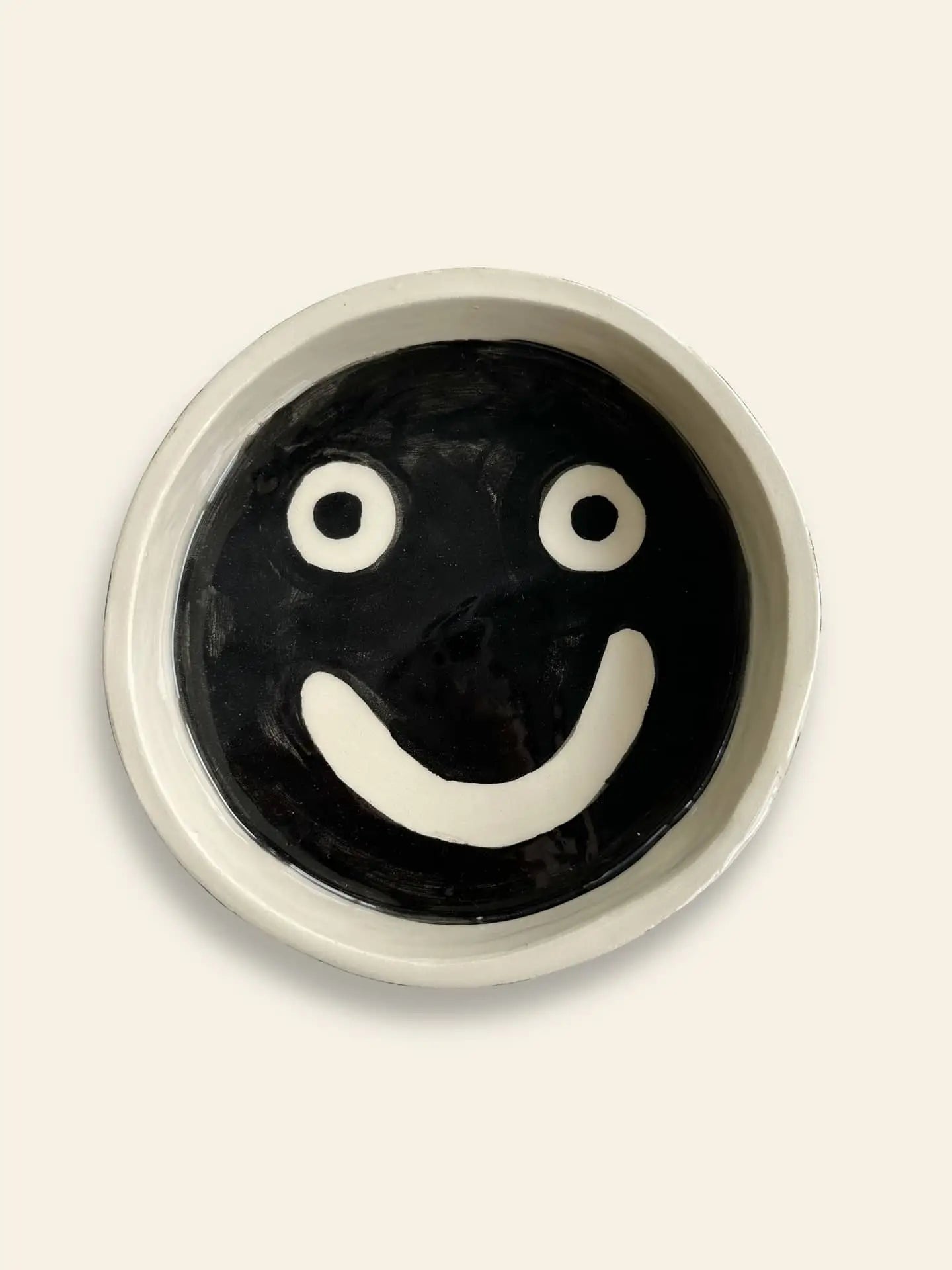 The Wobbly Studio Mood Tray is a round ceramic plate with a black background and a simple white smiling face. Ideal for personalized display or brightening up your space, this Wobbly Studio piece stands out on any light-colored surface.