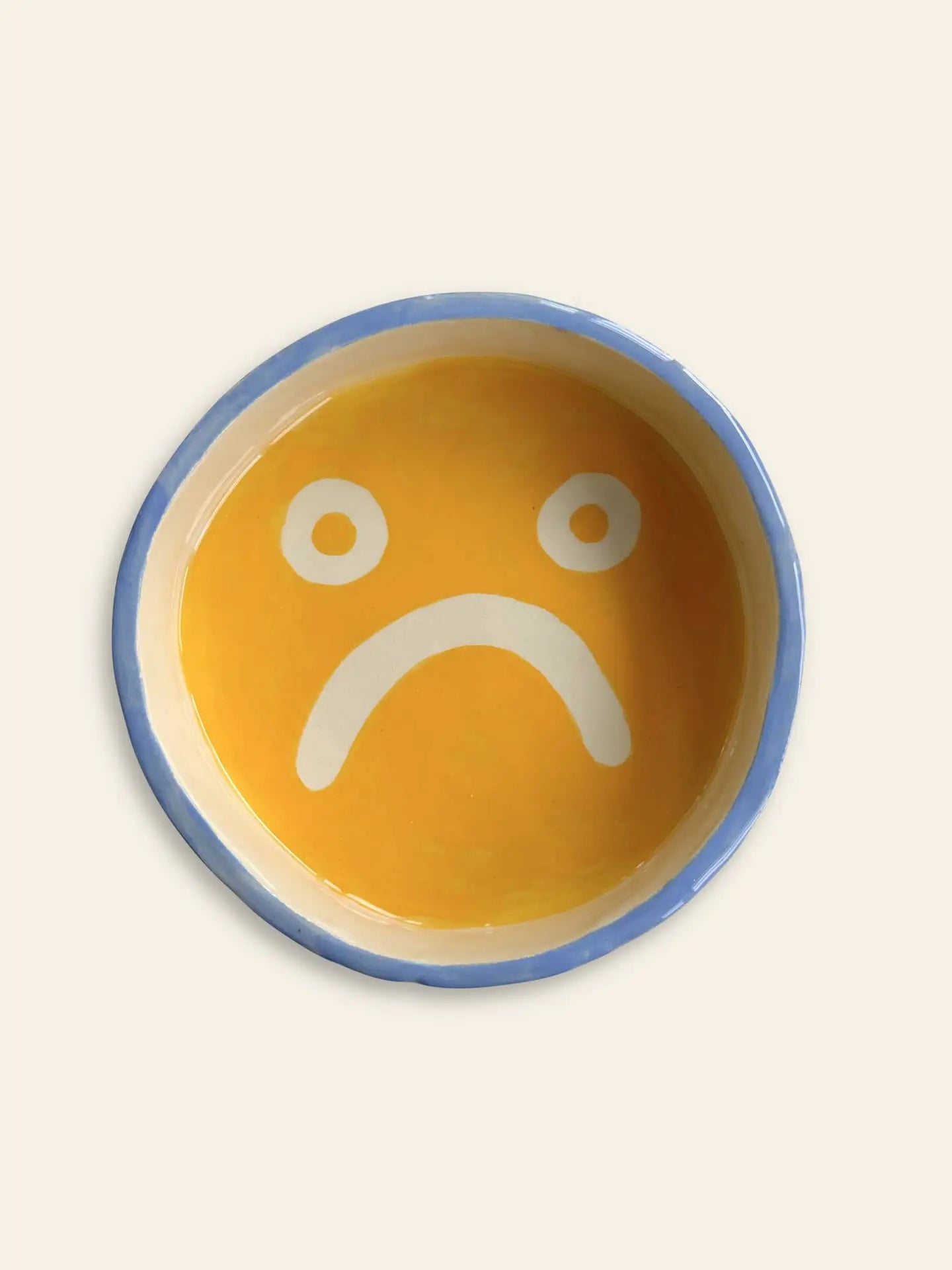 Brighten your space with the Wobbly Studio Mood Tray by Wobbly Studio—a round ceramic tray with a yellow center, blue rim, and a white sad face for a playful, personalized display.