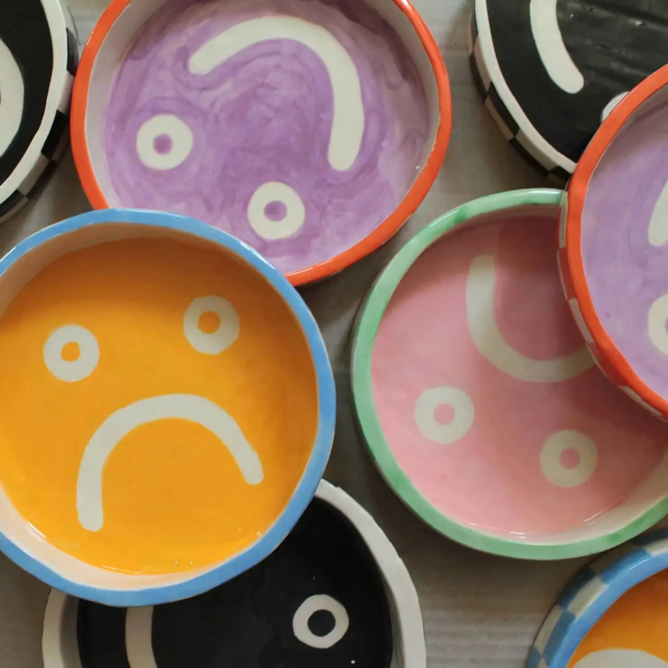 Wobbly Studio Mood Trays by Wobbly Studio are small, round plates in vibrant purple, orange, pink, black, and yellow shades with a white sad face, arranged overlapping for a playful personalized display.