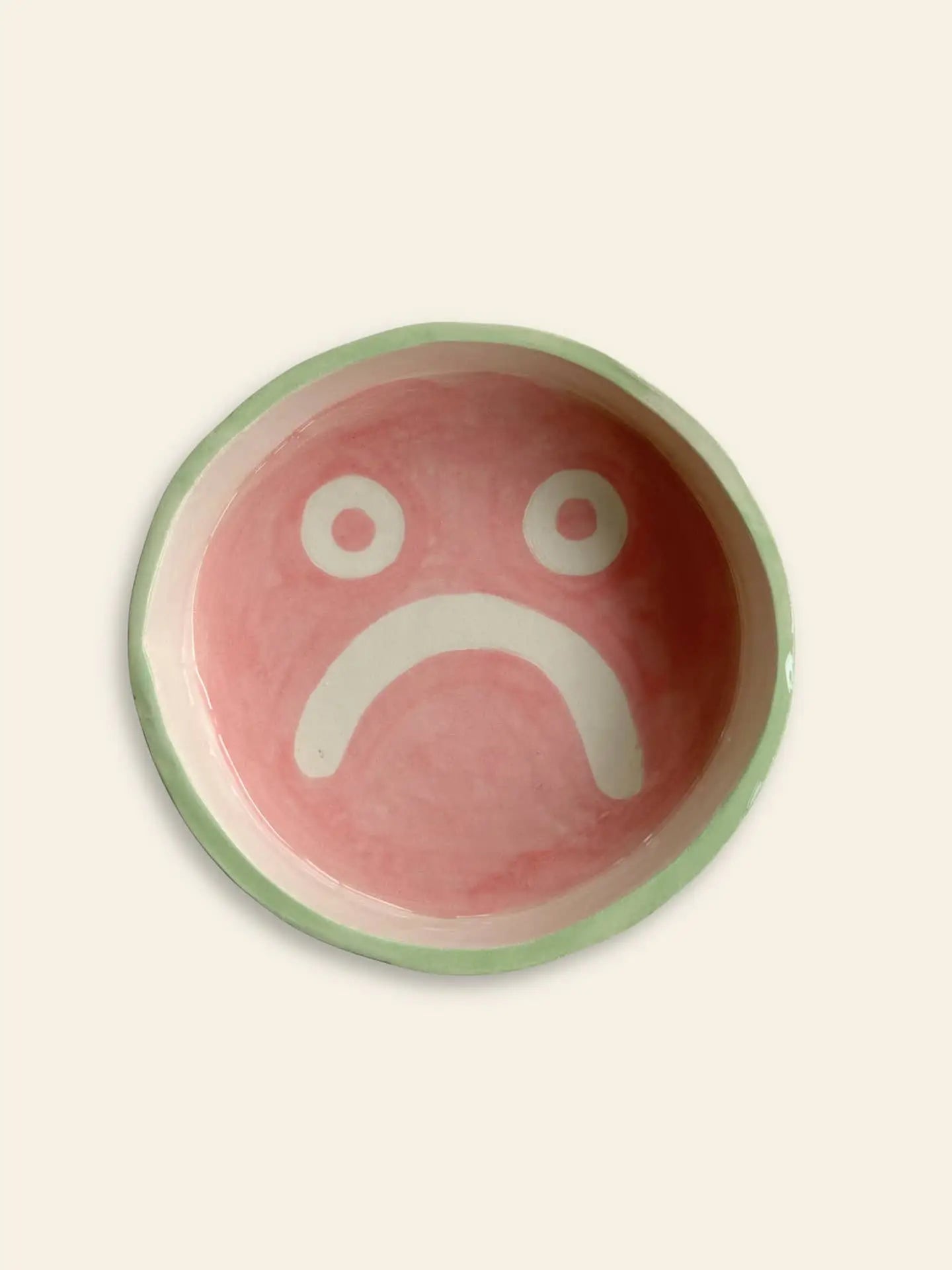 The Wobbly Studio Mood Tray by Wobbly Studio is a round ceramic plate with a pale green rim, pink center, and white sad face—its soft, vibrant colors add charm and personality to your display.