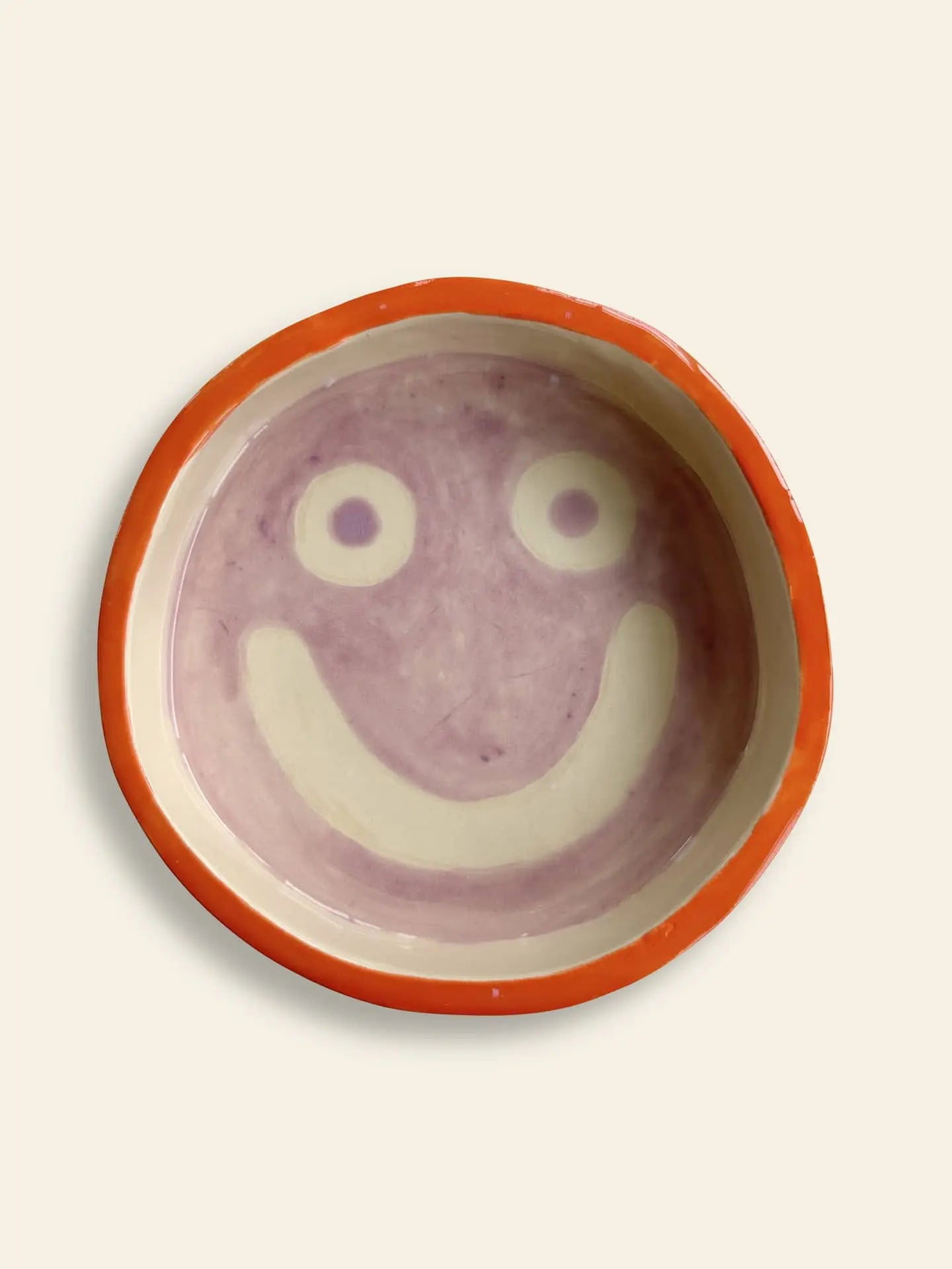 The Wobbly Studio Mood Tray features a round ceramic bowl with a red rim and a cheerful smiling face in light purple on cream, ideal for brightening up personalized displays.