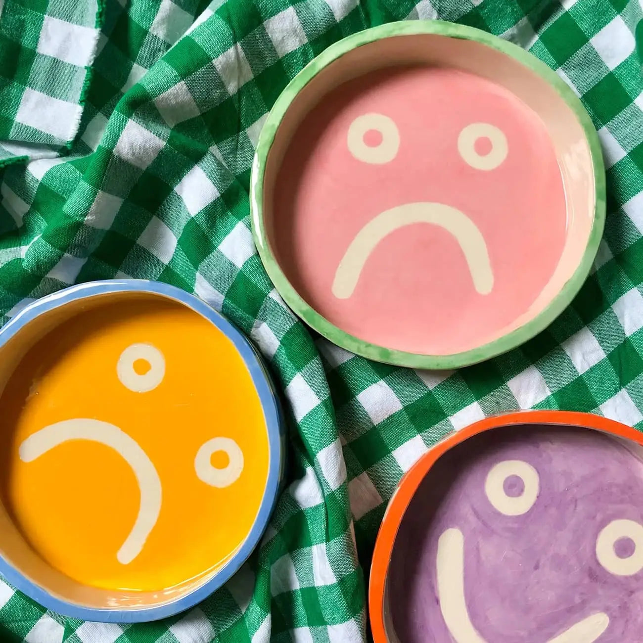 Add a splash of color to your décor with Wobbly Studio Mood Trays—three round trays in pink, yellow, and purple with sad face designs by Wobbly Studio—displayed on green and white checkered fabric.
