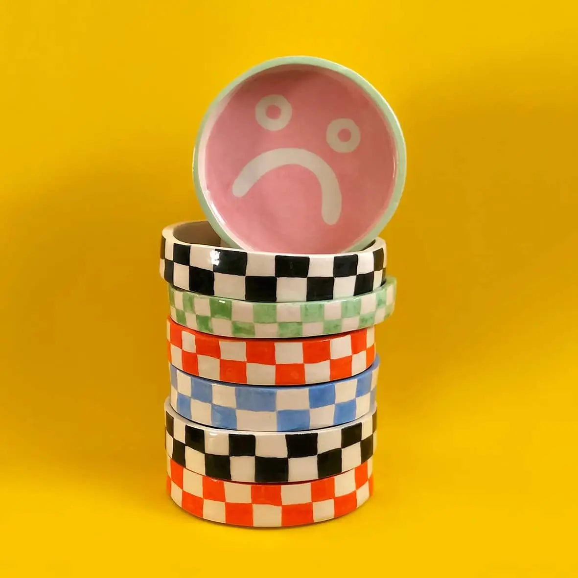 A stack of Wobbly Studio Mood Trays by Wobbly Studio, featuring vibrant checks, sits on a yellow background. The top tray has a pink interior with a large white sad face—ideal for adding personality to your display or tabletop.