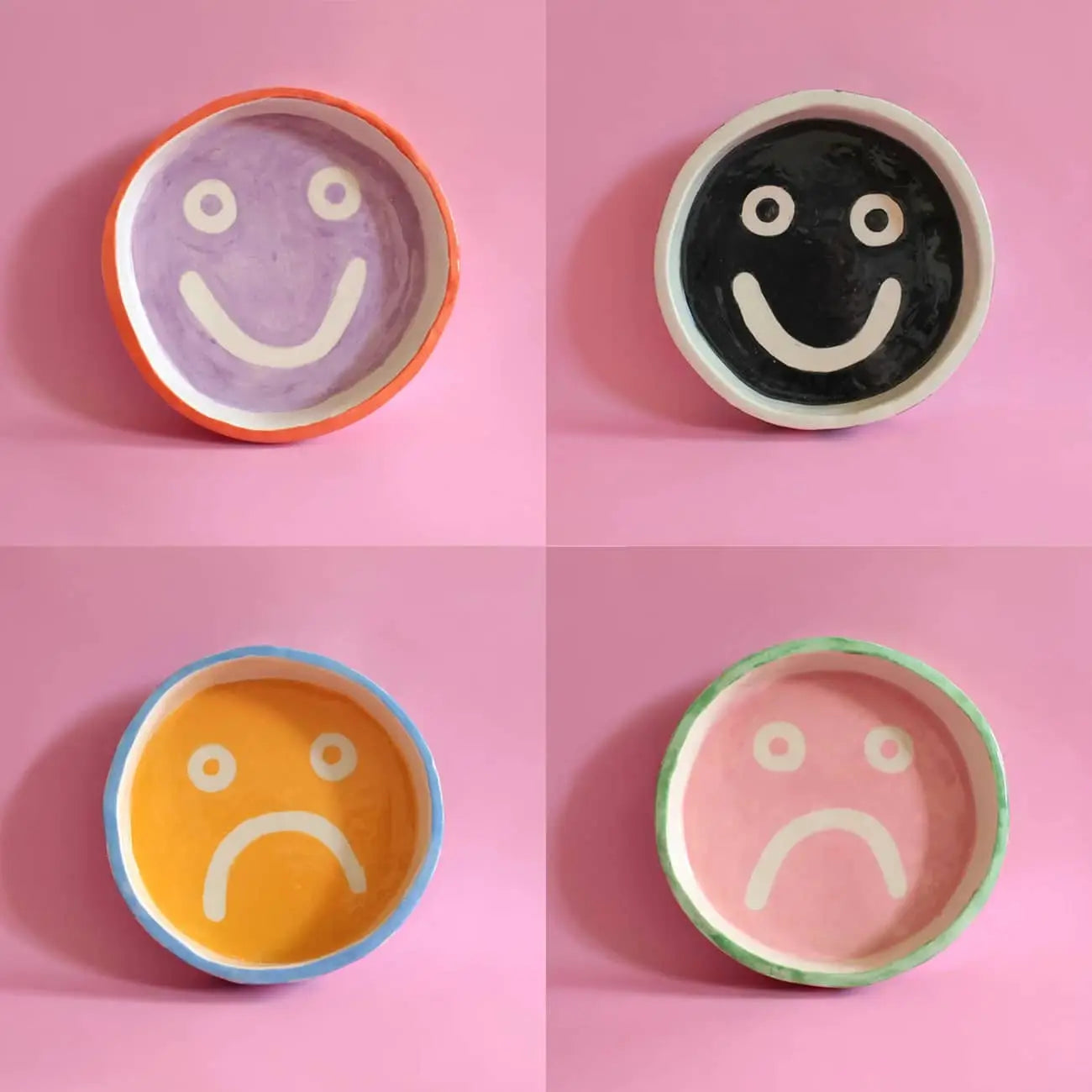 Four Wobbly Studio Mood Trays by Wobbly Studio are arranged on a pink background: purple (top left) and black (top right) trays have white happy faces, yellow (bottom left) and pink (bottom right) trays have white sad faces.