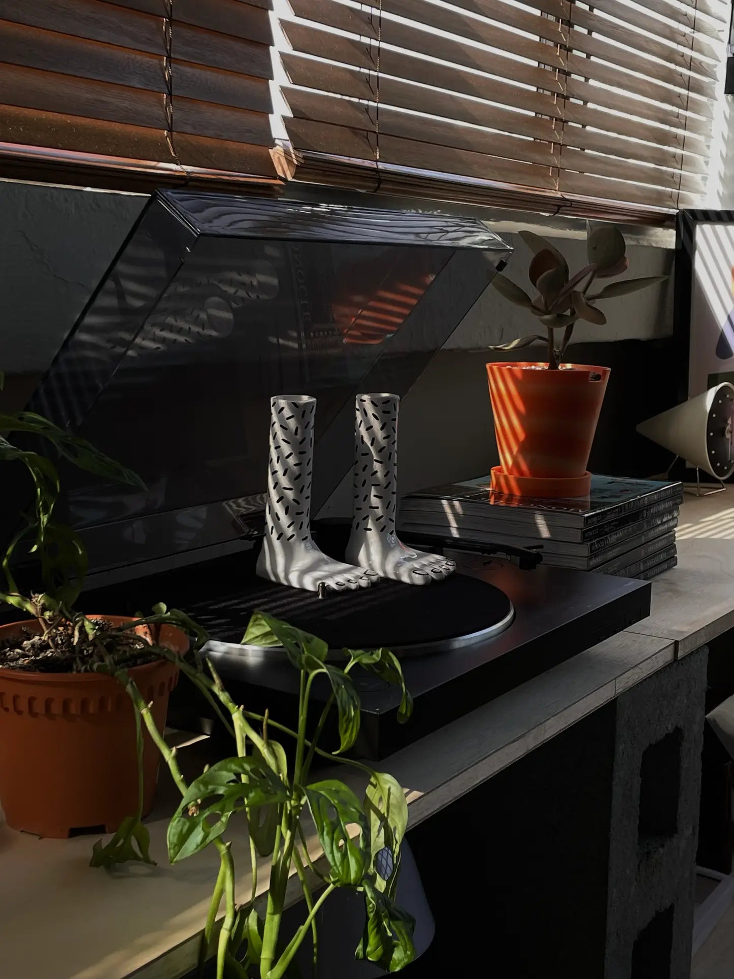 The Wobbly Studio Pair Of Legs Vase by Wobbly Studio, a white decorative boot-shaped piece with black patterns, rests on a record player amid potted plants, books, and sunlit window blinds.