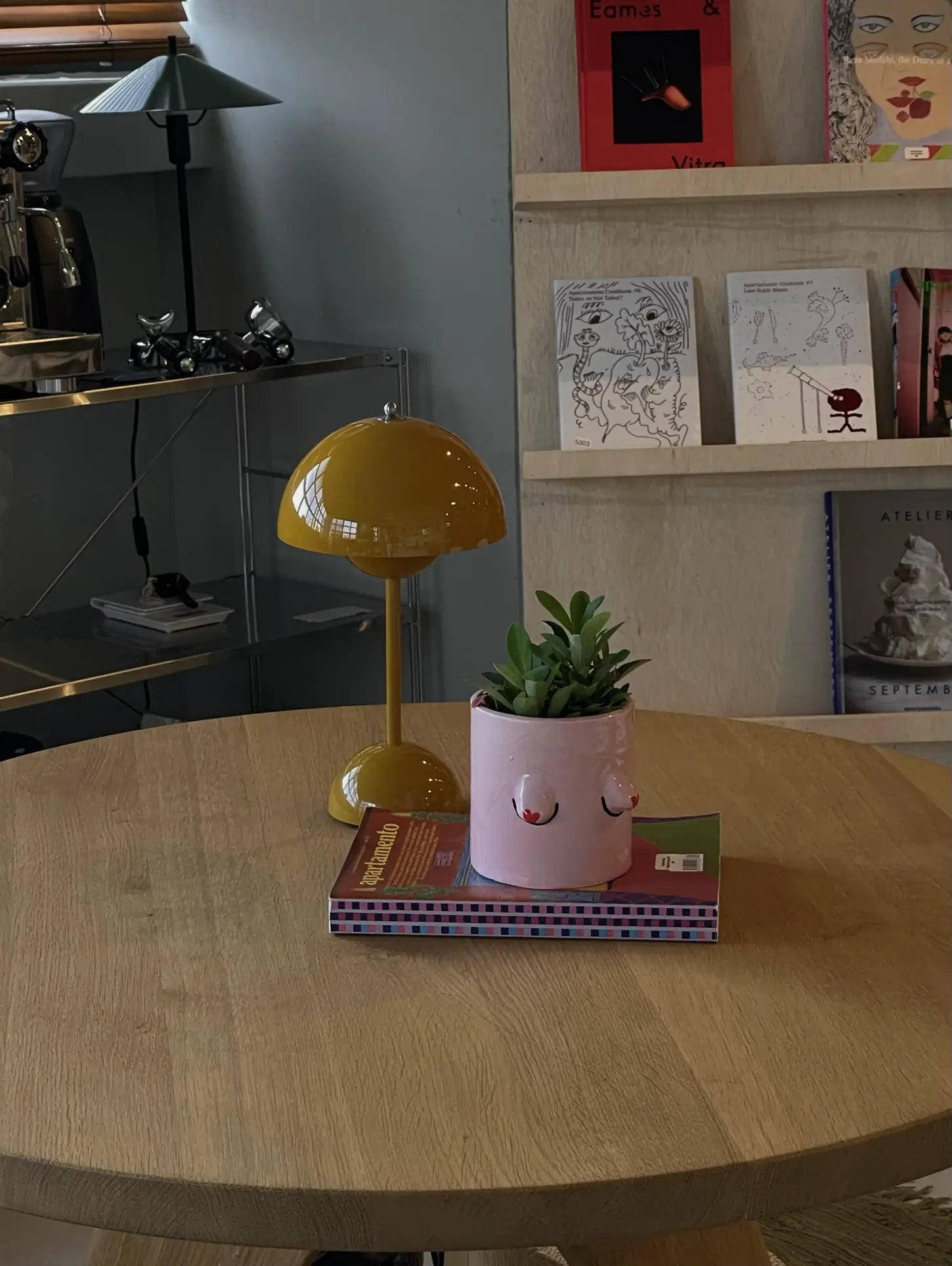 A round wooden table with a playful plant setup: a pink Wobbly Studio Regular Boobies Pot by Wobbly Studio, a green succulent, two books, and a yellow lamp. Shelves with books, art, and a metal shelf with a coffee machine are in the background.