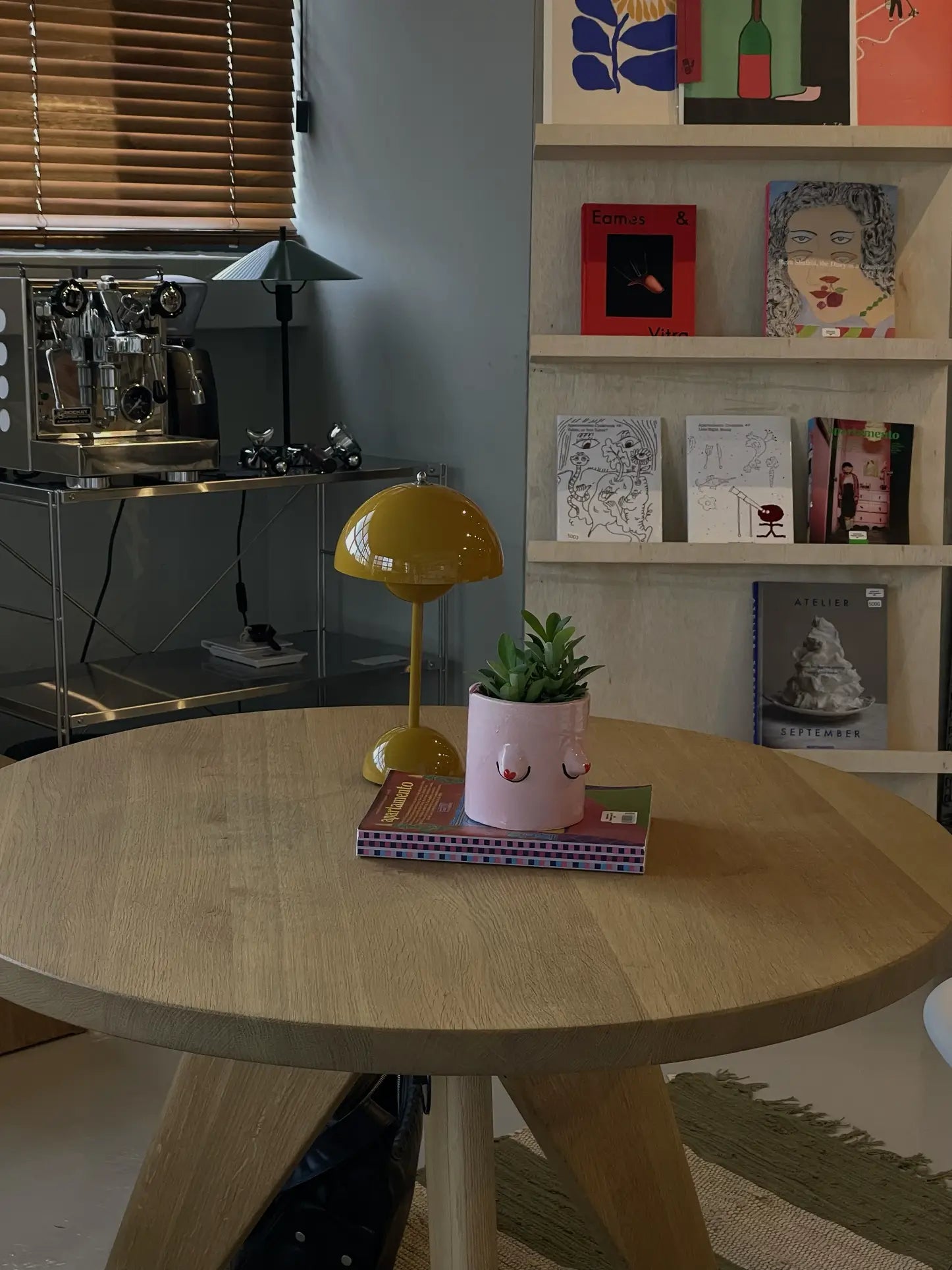 A round wooden table features a yellow lamp and a Wobbly Studio Regular Boobies Pot by Wobbly Studio with a succulent. Behind, shelves hold books and art, while a coffee machine sits on a metal counter by a window with brown blinds.