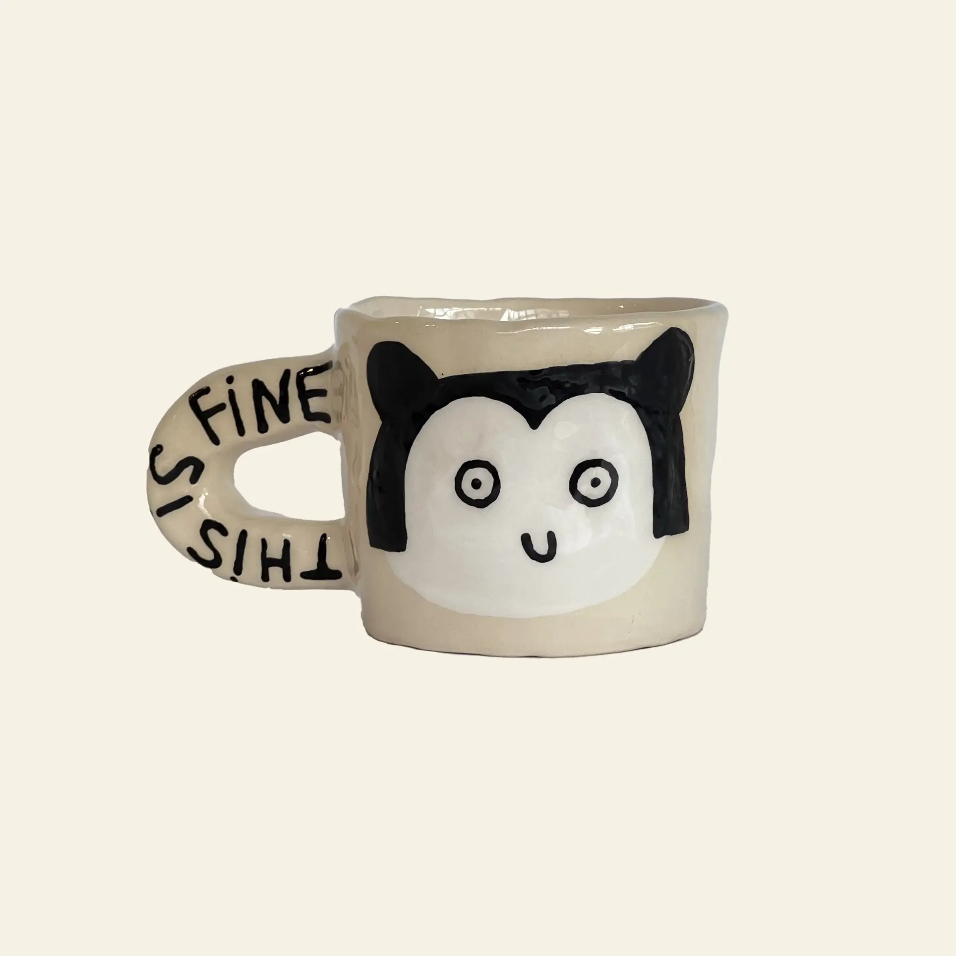 The Wobbly Studio This Is Fine Mug by Wobbly Studio features a beige, handcrafted design with a cartoon animal face and black ears. THIS IS FINE is printed on the handle, adding whimsy to any drinkware collection. Inspired by Marie Chanteur’s illustration.