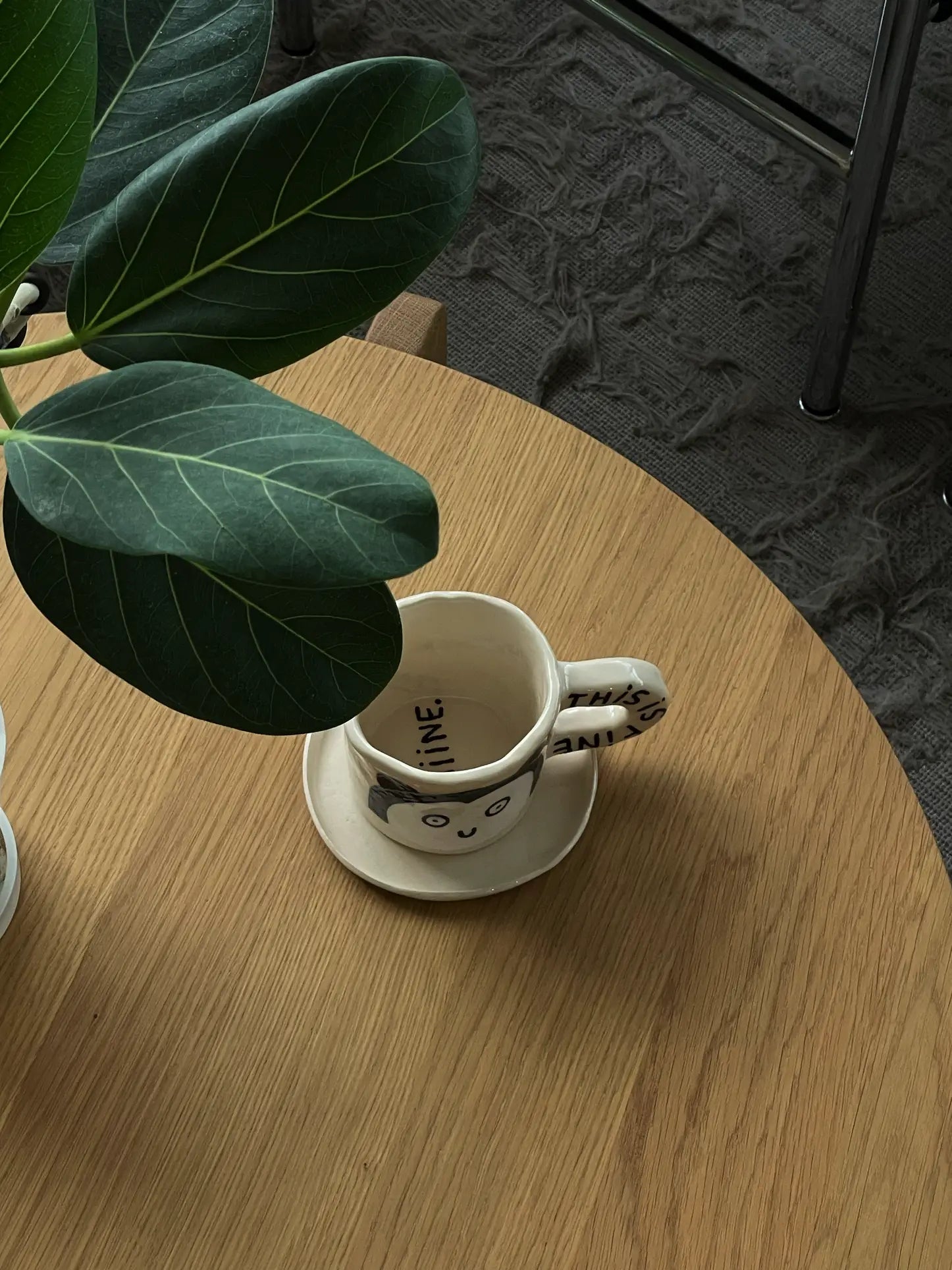 The Wobbly Studio This Is Fine Mug, featuring a cat face design, rests on a wooden table beside a leafy green plant, with part of a chair and textured carpet in the background.