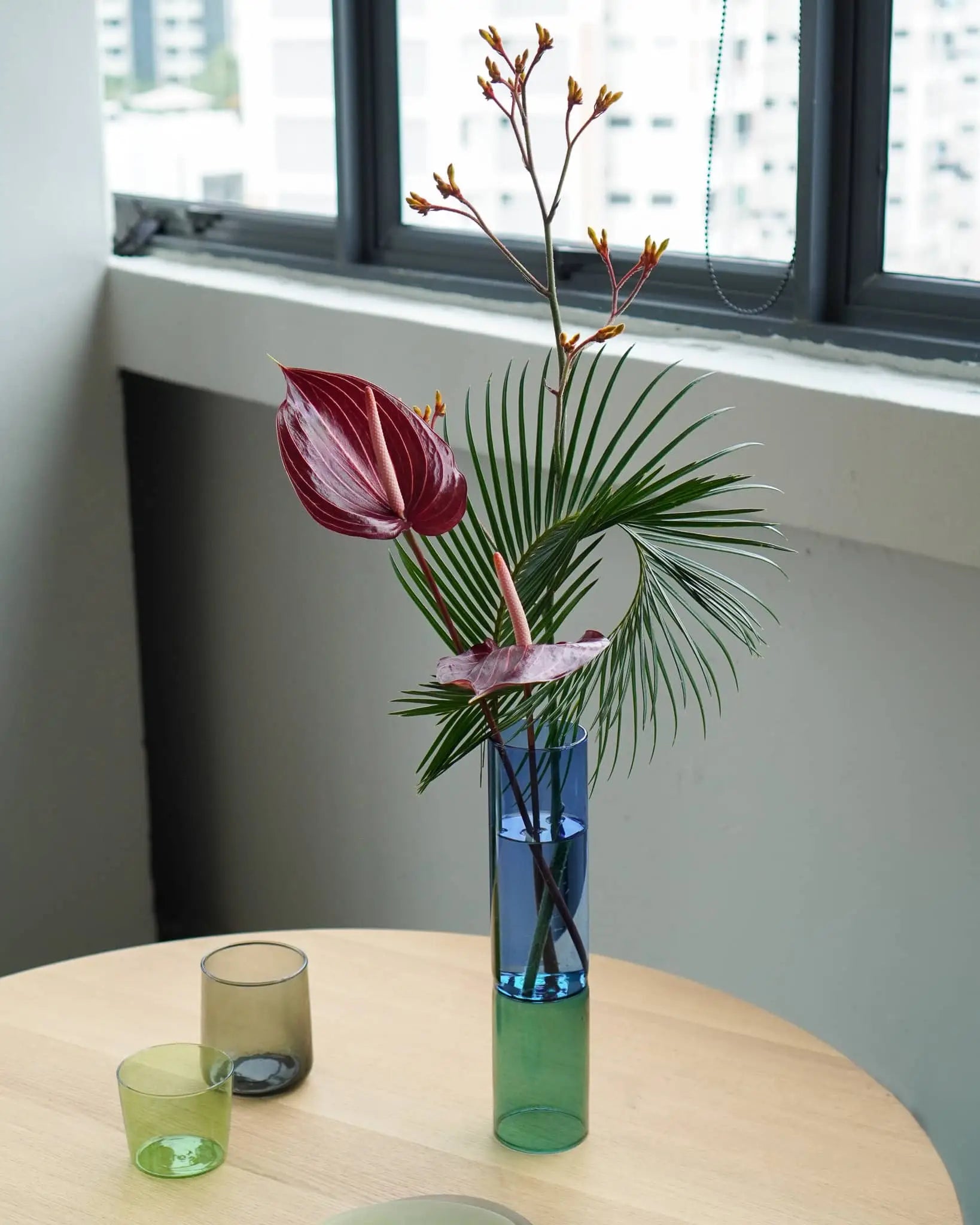 The Wonderland Botanicals Bamboo Groove Vase Arrangement by Wonderland Botanicals features a modern anthurium display with palm leaves and orange-tipped stems in a handmade blue-green vase on a wooden table, accompanied by glass cups and city views outside the window.