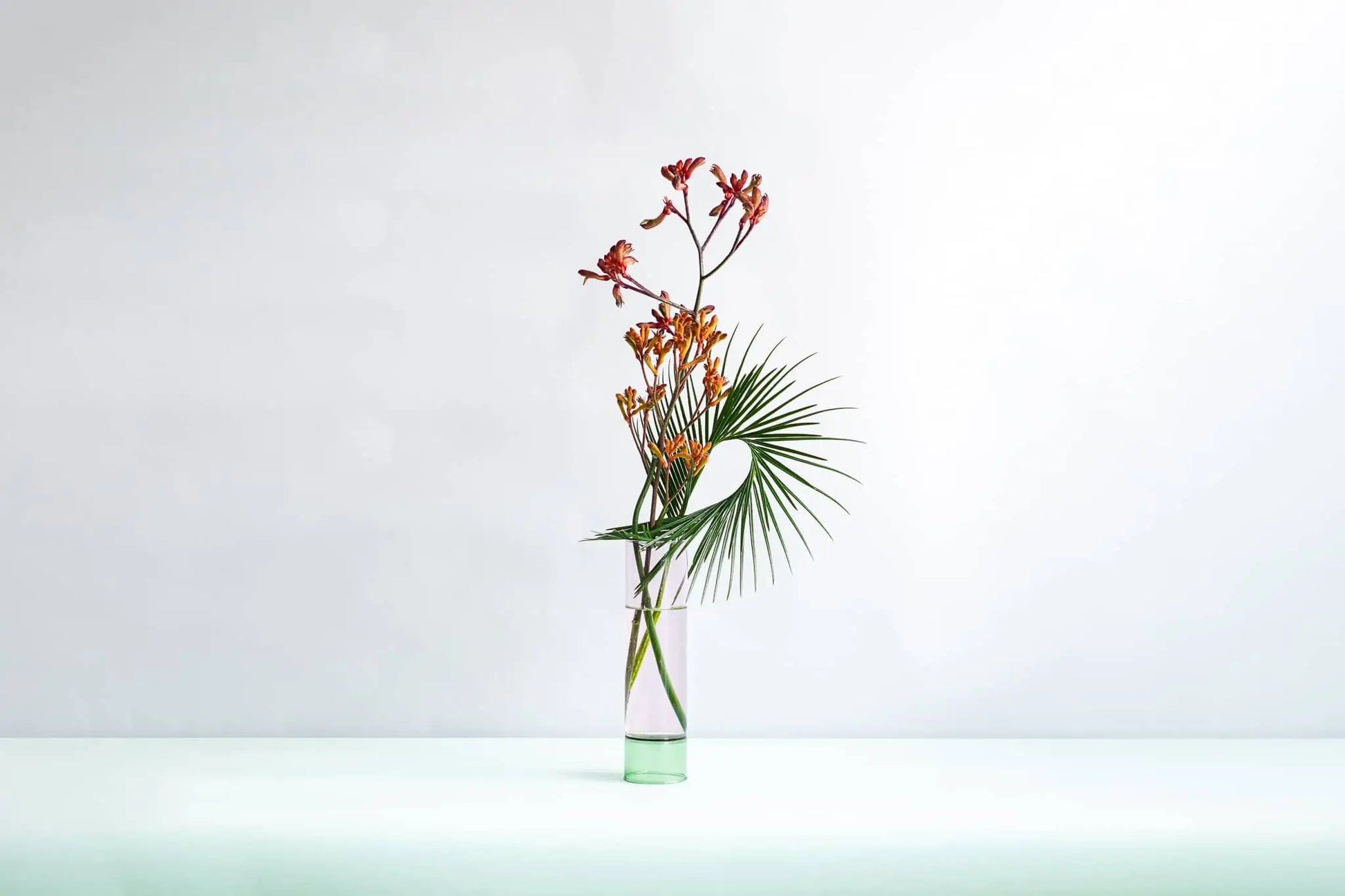 The Wonderland Botanicals Bamboo Groove Vase Arrangement features orange flowers and a green palm leaf, adding unique textures to your decor. Displayed on a light surface with a white background, in collaboration with Wonderland Botanicals.