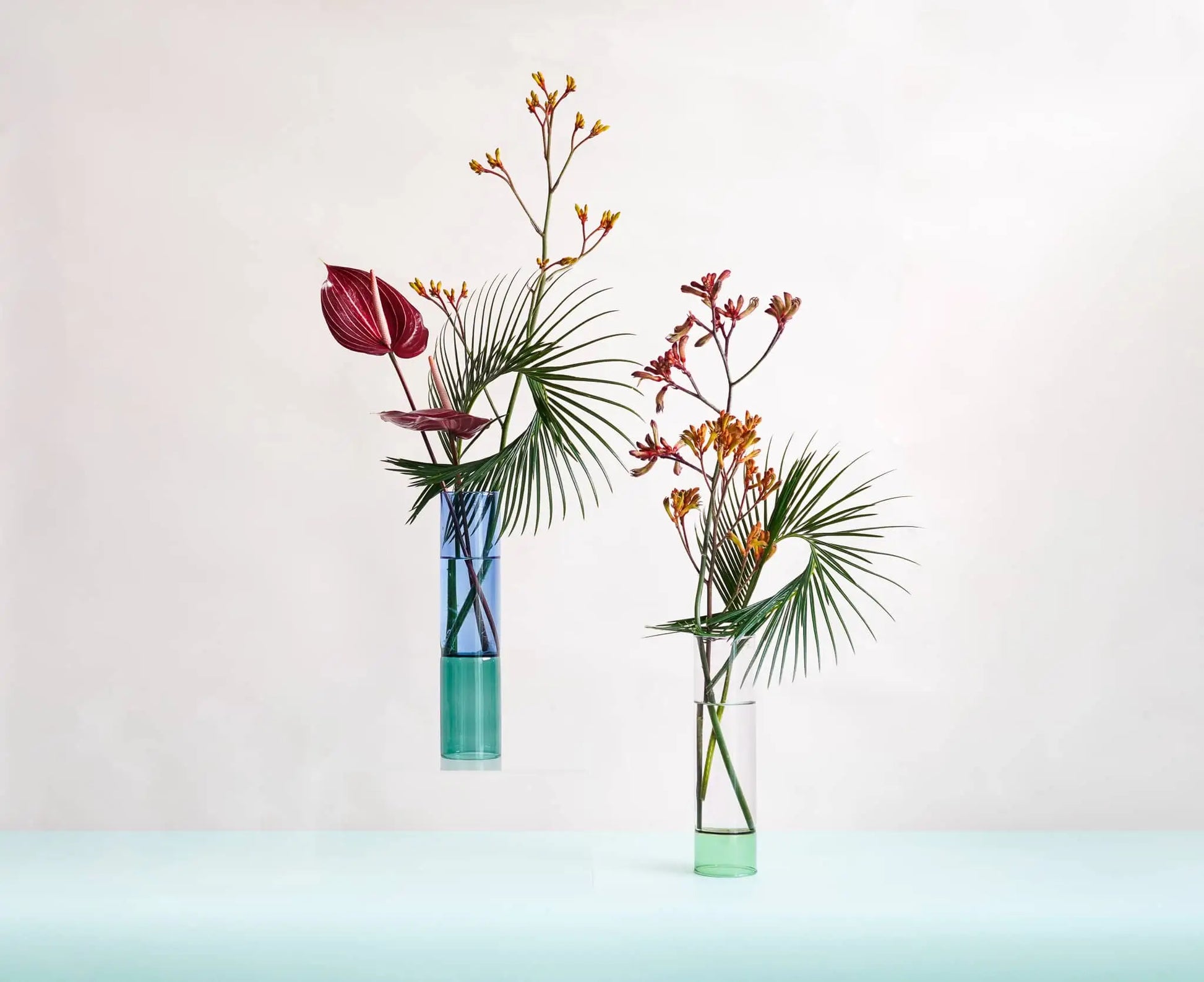 Two Wonderland Botanicals Bamboo Groove Vase Arrangements display palm leaves, red anthurium, and yellow kangaroo paw flowers in green water, adding vibrant color and texture to your home decor against a light background.