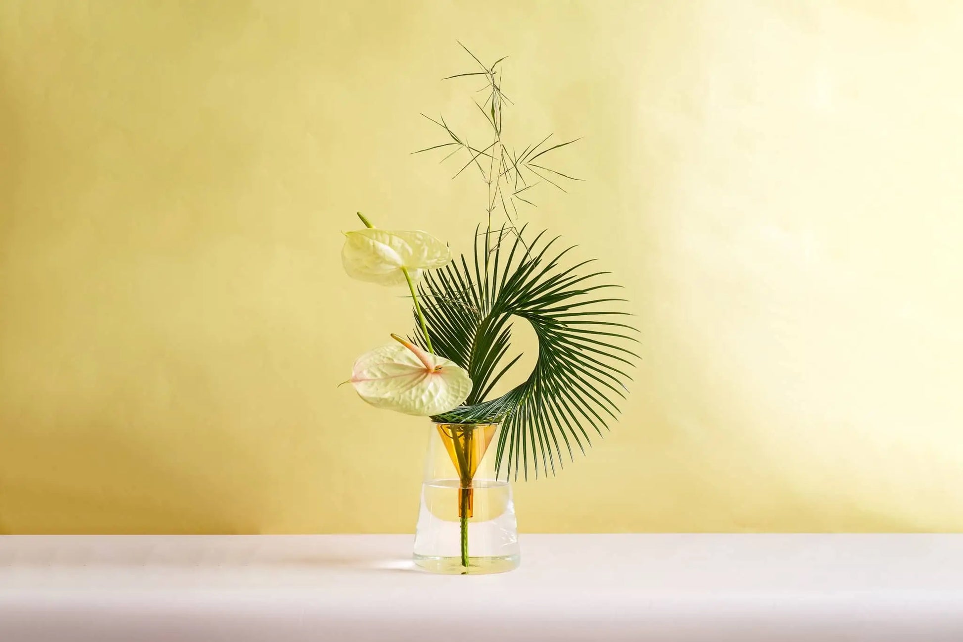 The Wonderland Botanicals Kado Vase Arrangement features palm leaves, bamboo foliage, and white anthurium in a clear vase with gold accents by Wonderland Botanicals, set against a light yellow background.