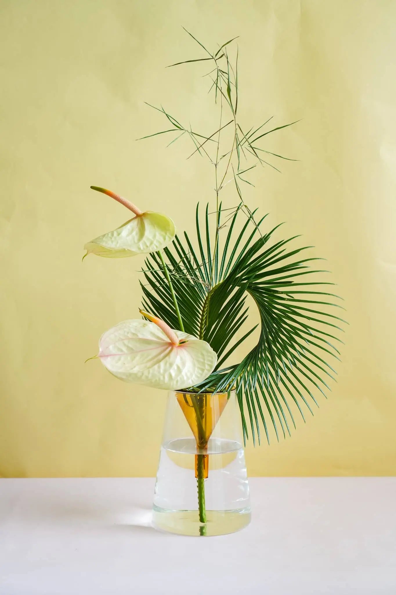 The Wonderland Botanicals Kado Vase Arrangement features two pale anthuriums, graceful bamboo foliage, and a large curved palm leaf in a glass vase by Wonderland Botanicals, set against a light yellow background.