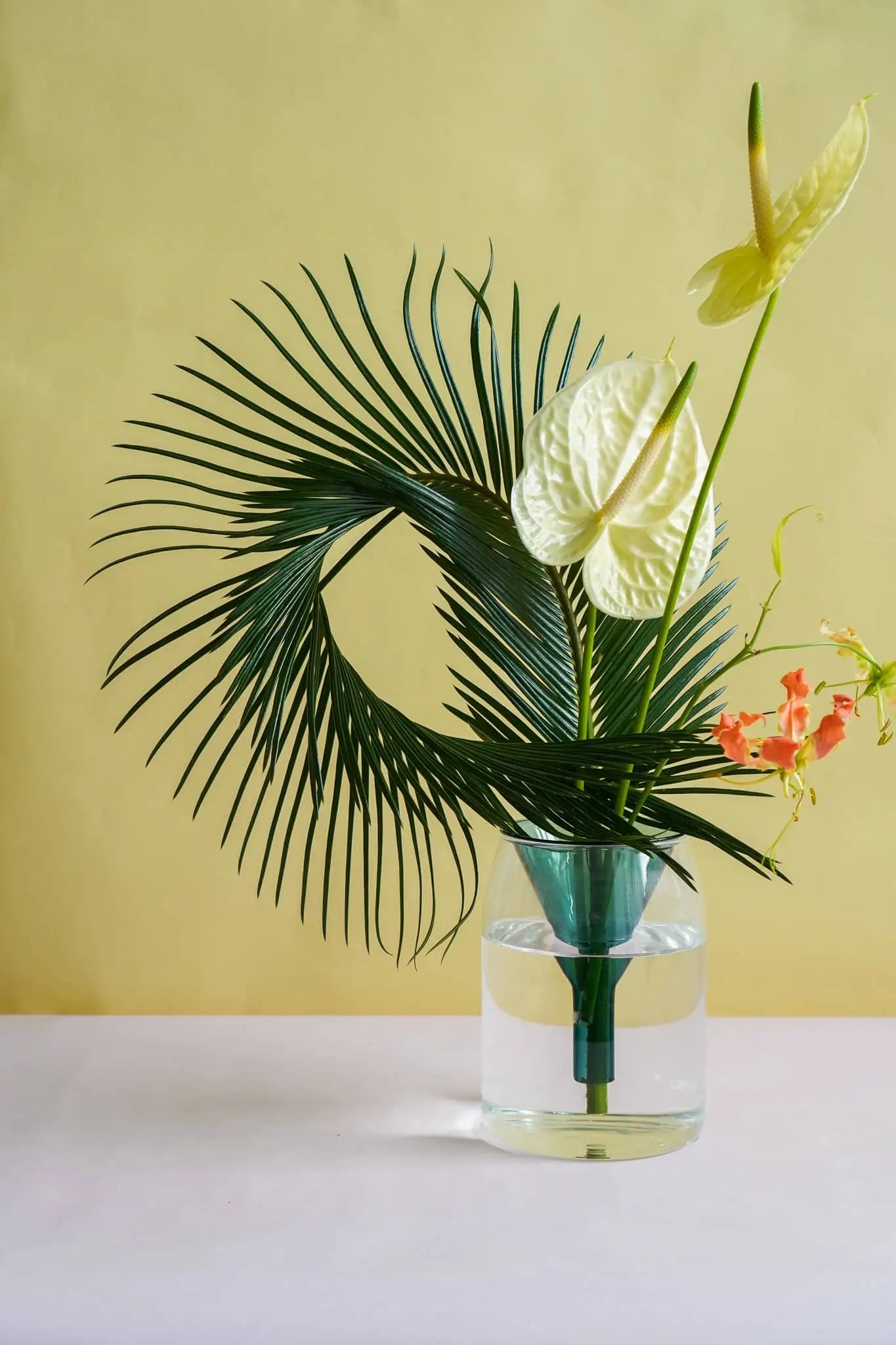 The Wonderland Botanicals Kado Vase Arrangement by Wonderland Botanicals features green palm leaves, pale yellow anthuriums, and orange flowers in a vase, set against a light yellow background on a pale surface.
