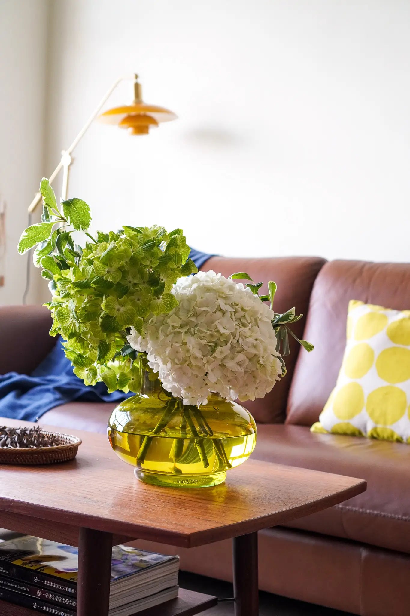 An Olive vase from the Wonderland Botanicals Collaboration holds white hydrangeas and green leaves on a wooden coffee table, set before a brown sofa with a yellow polka-dot pillow in this cozy, modern Wonderland Botanicals-inspired living room.