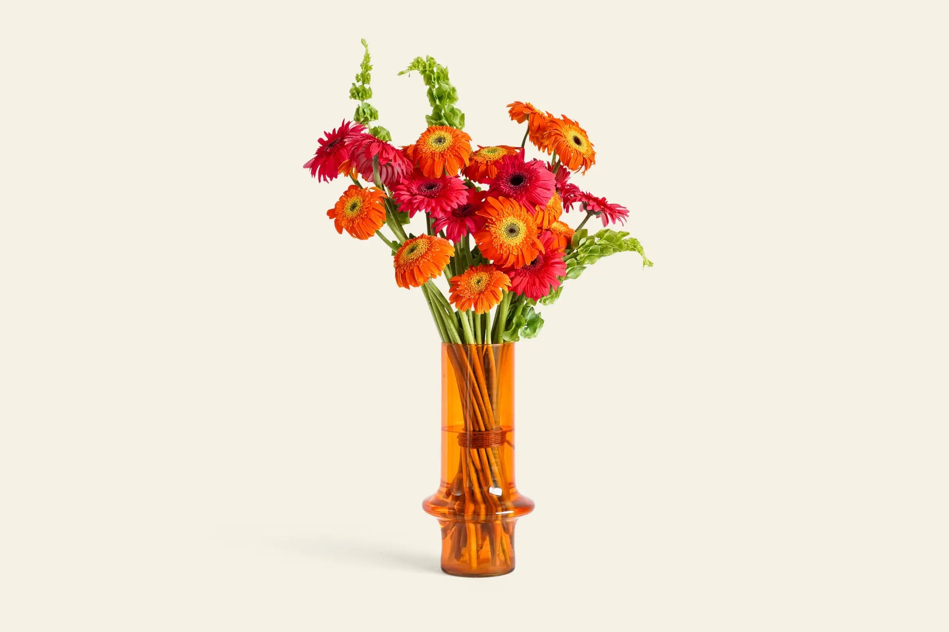 Paprika by Wonderland Botanicals is a bold, colorful floral arrangement featuring orange and pink gerbera daisies with green accents in a tall amber vase—a striking dining table showpiece from the Wonderland Botanicals collaboration.